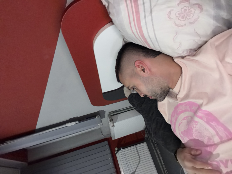 The scene is located in a room in Opanets, Bulgaria. An adult male is depicted lying on his back, with his head resting on a pillow, looking towards the left side of the frame. He has short dark hair with faded sides and a short beard. A black in-ear monitor is visible in his right ear. He is wearing a light pink short-sleeved t-shirt with a light pink graphic design on the front. A dark wristband is present on his left wrist. He is partially covered by a white duvet or blanket patterned with pink and grey floral motifs. The bed features a modern red and white headboard. In the background to the left, a window with grey horizontal blinds is visible, beneath which a white radiator is mounted. The wall exhibits a distinct design with an upper red section and a lower white section, divided by a diagonal line. A long, grey vertical fixture is mounted on the wall near the window. A dark grey textured fabric, possibly another blanket, is visible on the bed to the man's left.