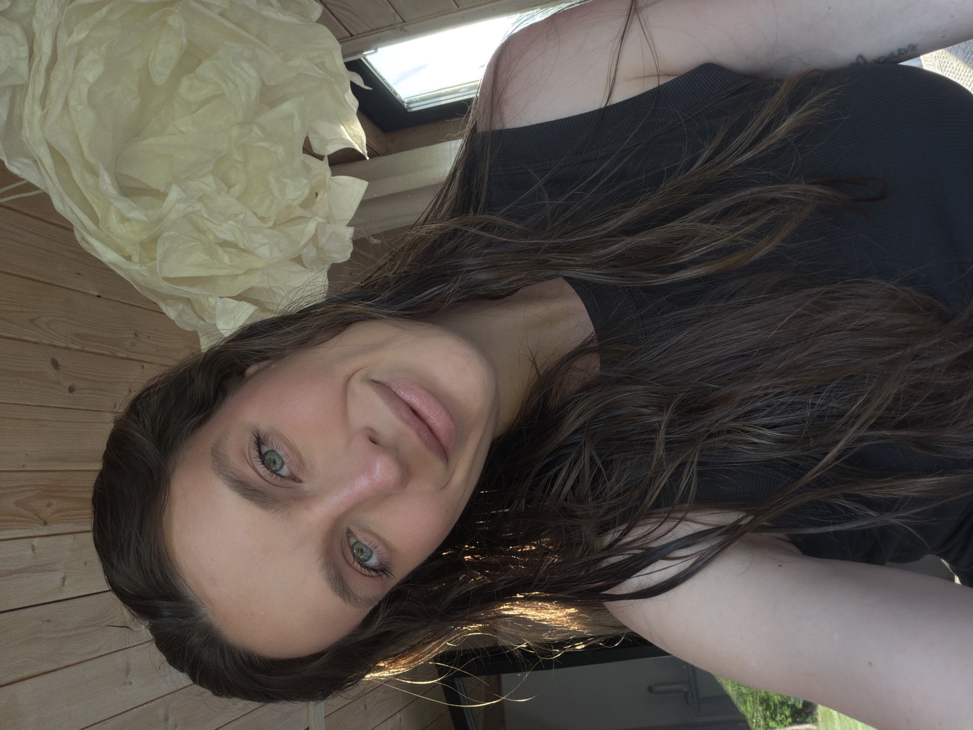 A young woman with long brown hair and green eyes is facing the camera. She has light skin and appears to be wearing a dark-colored top. She is smiling slightly. The image is slightly rotated.
The background includes wooden paneling and a crumpled white object resembling paper or fabric, possibly a decoration. There is also a skylight visible.
There is no information in the image to confirm its location in Prudki, Russia.