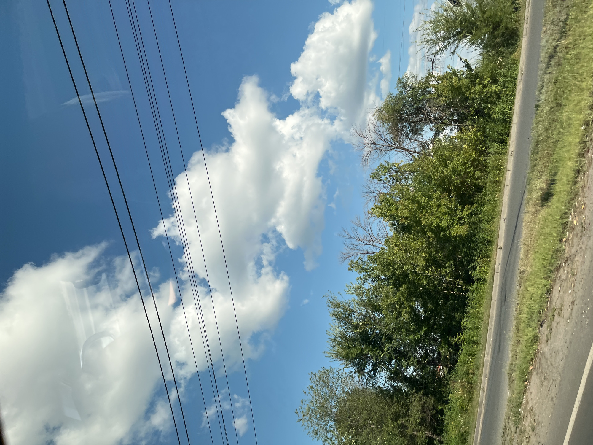 The image depicts a road cutting through a vegetated area under a bright blue sky dotted with fluffy white clouds. A line of trees and bushes borders the road on the right side. Several power lines are strung horizontally across the upper portion of the frame. The image appears to have been taken from a moving vehicle, possibly through a window, as evidenced by reflections on the sky and clouds. The scene is devoid of people and any specific landmarks that would identify it as being in Saratov, Russia. The environment suggests an urban or suburban fringe, possibly an industrial or undeveloped area adjacent to a road.