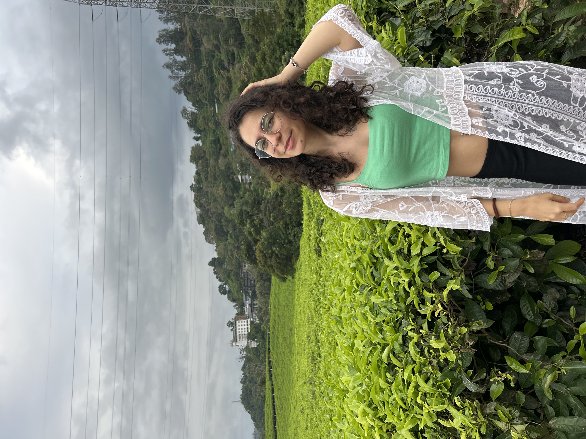A young woman is captured posing in a lush green tea plantation under an overcast sky. She stands on the right side of the frame, looking towards the viewer with a gentle smile. She has dark, curly hair and wears round-framed glasses. Her attire consists of a light green crop top, black pants, and a sheer, white lace long cardigan. Her left hand is casually raised to her head, touching her hair. A bracelet is visible on her left wrist.
The tea bushes, vibrant and dense, fill the foreground and midground, extending across gently sloping hills. In the distant background, a multi-story white building is nestled amidst the greenery on a faraway hillside. The sky above is completely covered with grey clouds, indicating overcast weather conditions. Several prominent power lines stretch horizontally across the upper left portion of the image, with part of a large transmission tower visible at the far left edge. The overall setting is serene and natural, consistent with a tea plantation in a hilly region like Pallivasal, India. There is no visible text.