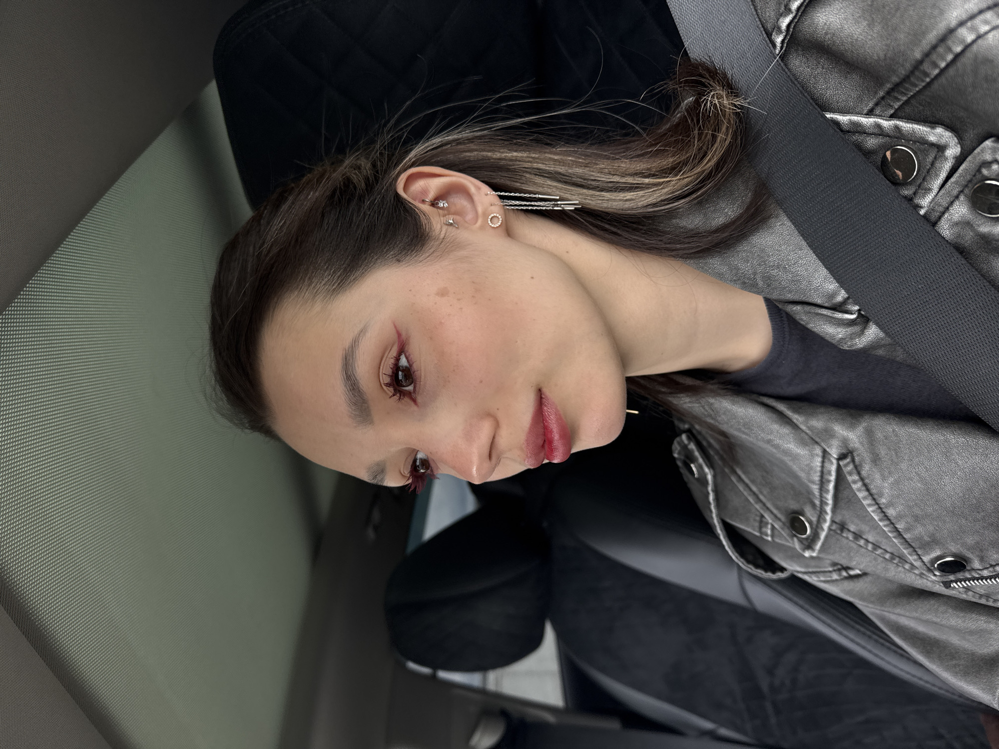 A young woman is seated in the passenger seat of a vehicle, likely a car, as indicated by the seatbelt across her chest and the dashboard/windshield visible in the background. She is wearing a grey leather jacket over a dark-colored shirt. Her hair is styled with lighter highlights. She has noticeable ear piercings, including multiple studs and a dangling chain earring. She has red makeup around her eyes and on her lips. The scene appears to be indoors, inside the vehicle. The location in the city of Unknown, Unknown, is not discernible from the image.