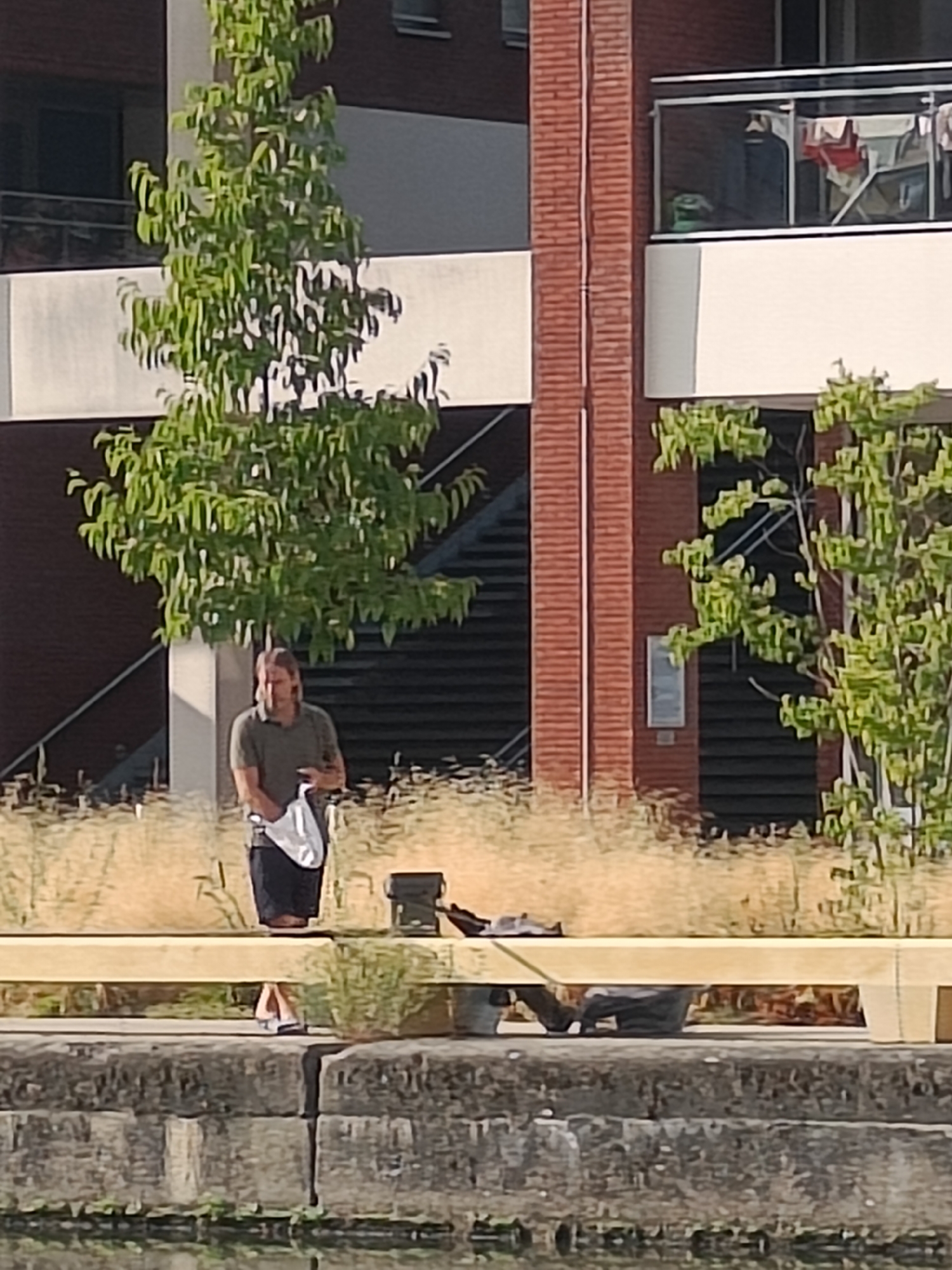 A man stands on a concrete structure near a body of water. He is wearing a dark green polo shirt and dark shorts, and he is holding a white bag. To his right, on the same concrete structure, is a black container. Behind him, a large tree with green foliage is visible, and further in the background is a brick building with balconies. The scene appears to be in an urban setting. The image was taken in Leuven, Belgium, near the Vaartkom.