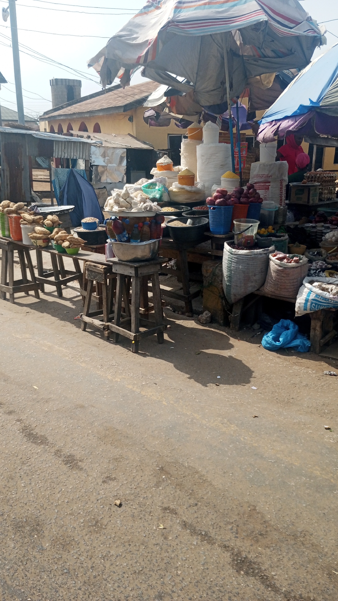 The image depicts a street market in Lokoja, Nigeria. Stalls line the roadside, displaying various produce and goods on wooden tables and stools. Items include potatoes, onions, grains, and flours. Vendors' merchandise is arranged under large umbrellas and awnings for shade. In the background, buildings with corrugated metal roofs and a distinct tower-like structure can be seen. The street is paved, with noticeable wear and tear. A blue plastic bag is visible near one of the stalls. The scene appears to be during daylight hours.