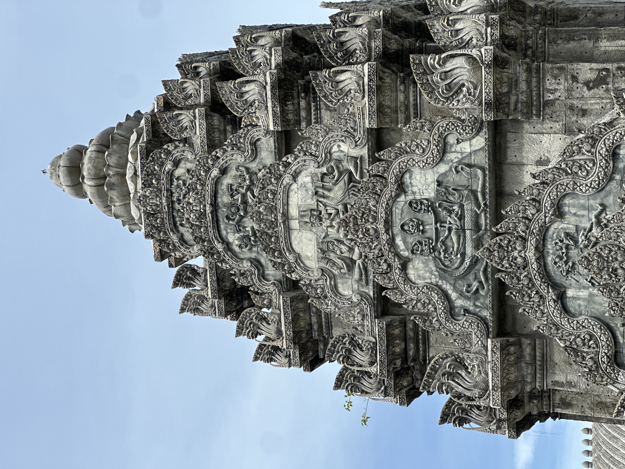 The image shows a close-up of an ornate, multi-tiered stone structure, likely a temple or monument, against a clear blue sky. The structure is heavily decorated with intricate carvings that appear to depict mythical figures, serpents, and decorative patterns. The stonework is weathered, suggesting age or exposure to the elements. The focus is on the detailed craftsmanship of the architecture, with multiple layers of tiered roofs and elaborate ornamentation. The composition is centered on the upper portion of the structure, emphasizing its height and detailed design.