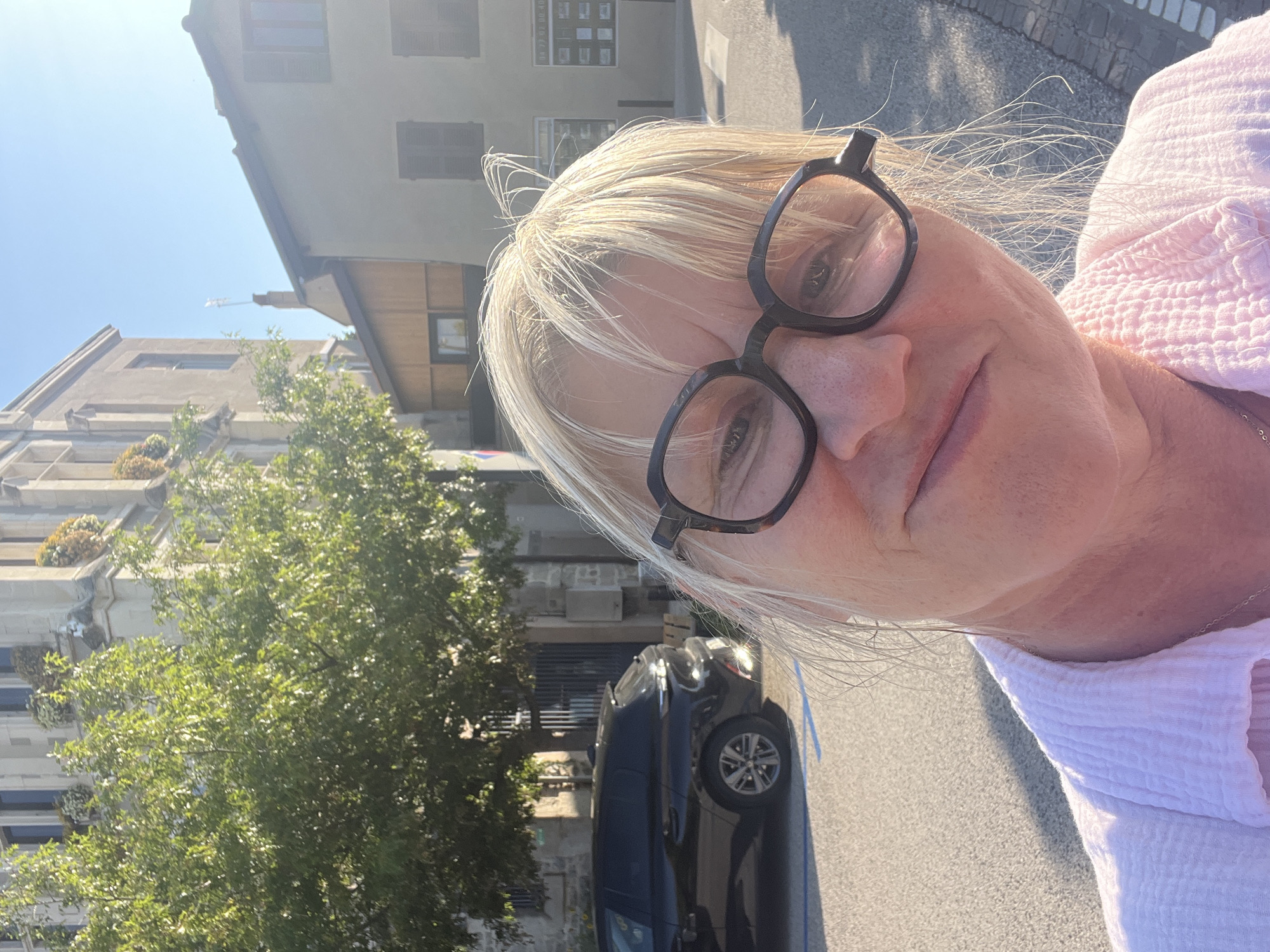 A woman with blonde hair and glasses is taking a selfie in front of a building in Saint-Galmier, France. She is wearing a pink shirt. A black car is parked on the street behind her, and a tree is to the left of the frame. The sky is blue and clear. The building appears to be an older, stone structure with multiple windows.