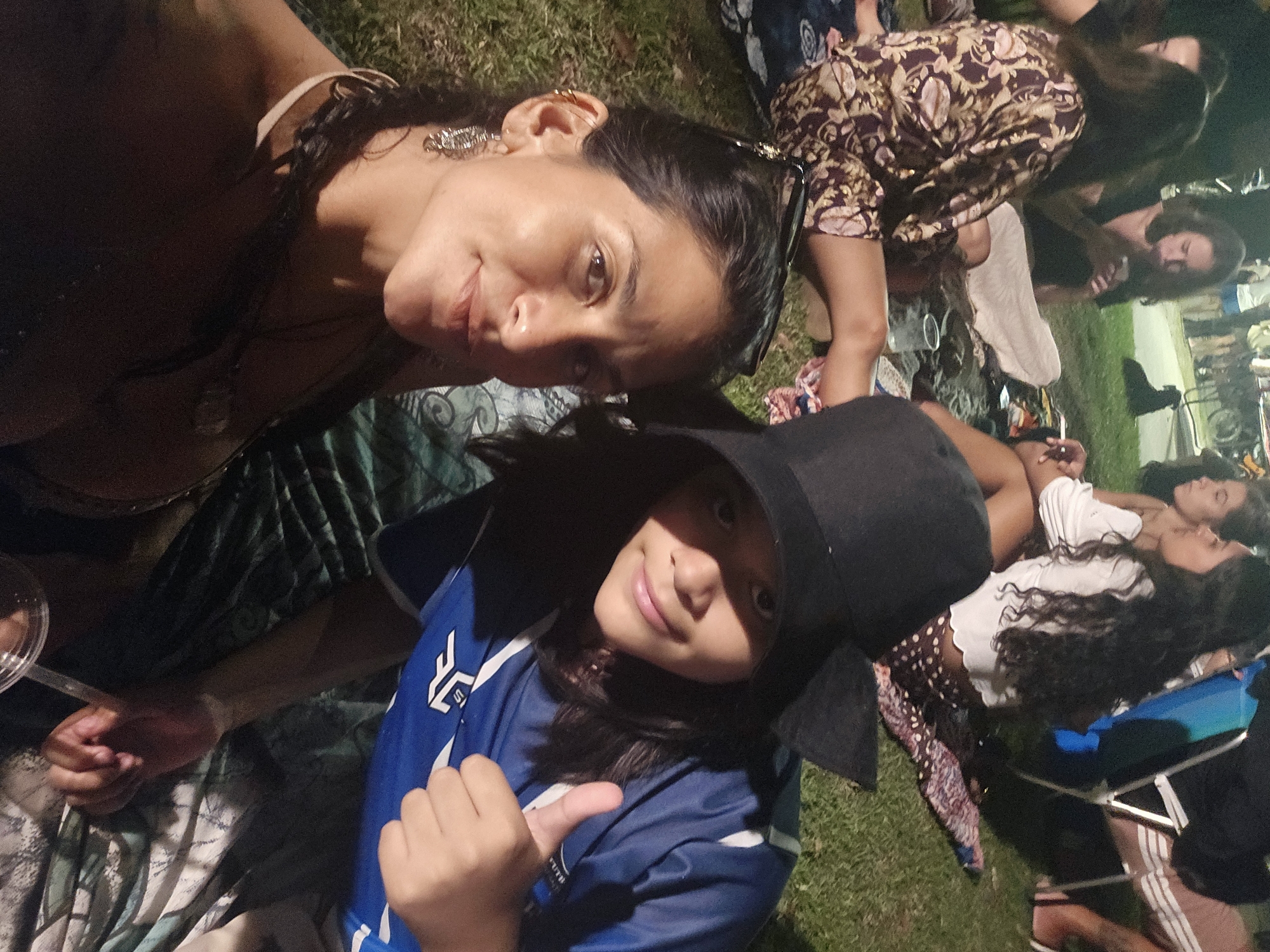 The image depicts a group of people gathered outdoors, likely at an event or gathering, in Armação dos Búzios, Brazil. A woman is taking a selfie with a child wearing a blue shirt and a black baseball cap who is giving a thumbs-up. Other people are seated on the grass in the background, and picnic items like blankets and drinks are visible. The environment seems relaxed and informal.