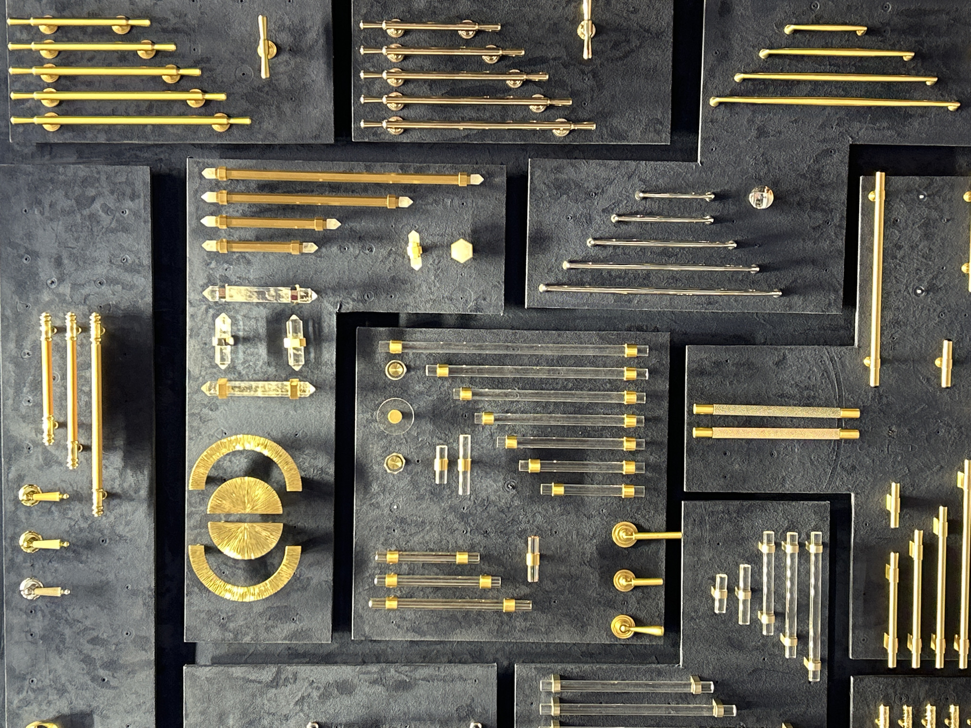 This image presents a detailed view of a diverse collection of cabinet hardware, including pulls, handles, and knobs, meticulously displayed on a series of dark, textured rectangular panels. The setting appears to be a showroom or design studio, showcasing various product options.
The hardware is predominantly metallic, featuring numerous gold or brass-colored bar pulls of different lengths and styles, some with smooth finishes and others with a textured, almost sand-blasted appearance. Silver or nickel-colored bar pulls are also part of the display, offering a cooler metallic alternative. A significant portion of the collection incorporates transparent elements, such as clear acrylic or lucite bar pulls with gold or brass accents, and pulls or knobs made with clear crystal or quartz, some faceted and others forming elongated clear bars.
Among the more distinctive designs are gold-colored half-circle pulls, several of which exhibit a radiating, textured pattern. There are also gold-toned T-bar pulls and door lever handles. The display is well-organized, with similar types of hardware often grouped together, highlighting the range of sizes, materials, and designs available. The dark, matte background effectively contrasts with the reflective metallic and transparent hardware, making each piece visually prominent. No people are visible, and there is no text present in the image. The scene is static, focused entirely on presenting the aesthetic and material variety of the hardware collection.