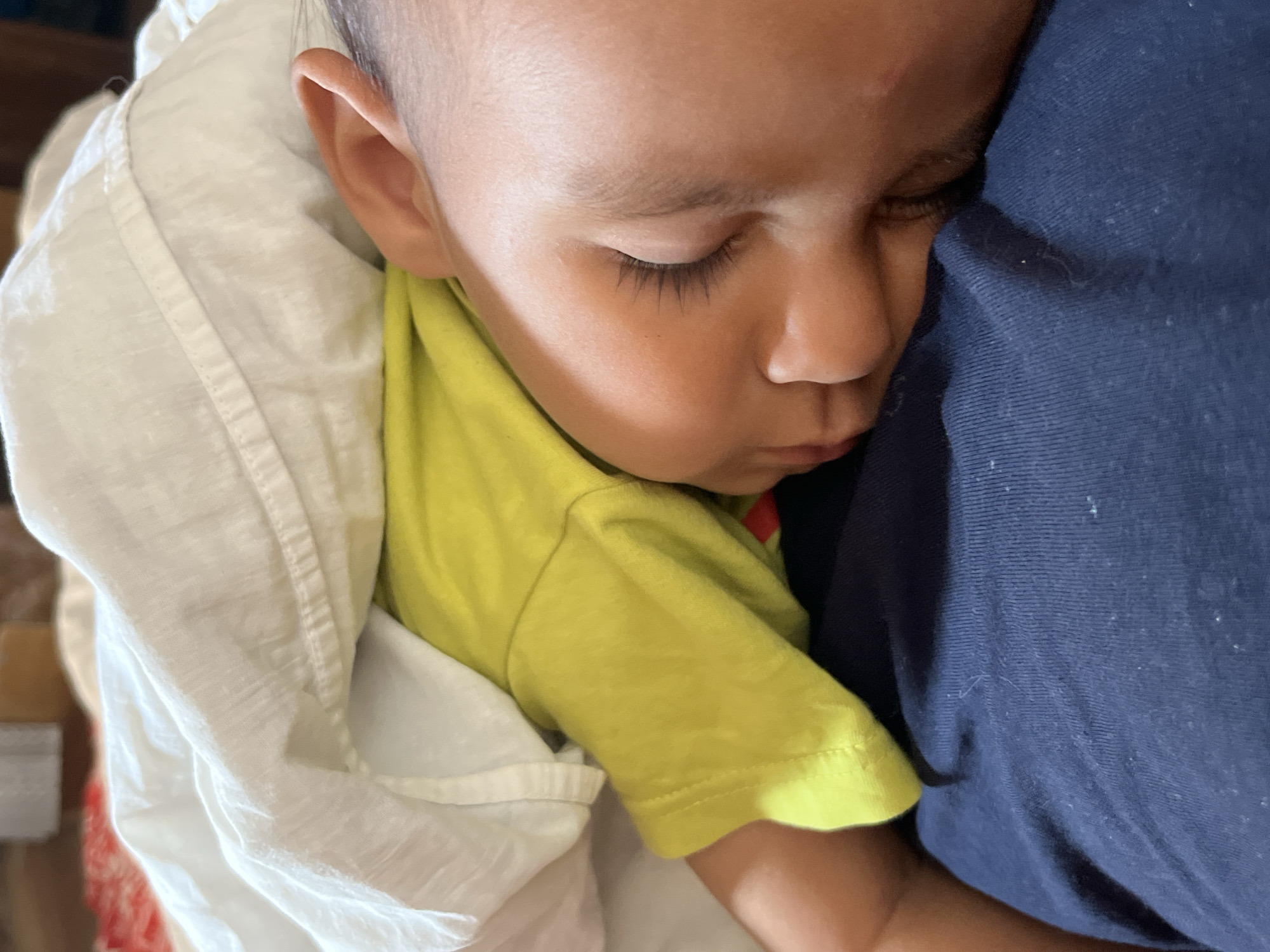 The image displays a close-up, high-angle view of a young child, likely a toddler, sleeping peacefully. The child is shown in profile, with their head resting gently against a dark blue fabric, which appears to be the clothing of another person, possibly a parent or caregiver, or a soft cushion. Their eyes are closed, revealing distinct, dark eyelashes, and their relaxed facial expression, with slightly parted lips, conveys a deep state of repose and comfort. A small, subtle red mark is visible on the child's forehead.
The child is dressed in a vibrant, lime-green short-sleeved t-shirt. A soft, white fabric, possibly a blanket or swaddling, is wrapped around their upper body and left arm, suggesting they are nestled and secure. The dark blue fabric against which the child is leaning displays visible texture and slight lint.
The overall scene evokes feelings of peace, warmth, and innocence. There are no discernible background details within the image to indicate a specific location in Oktyabrsky, Russia, or to suggest the time of day. No text is visible in the photograph.