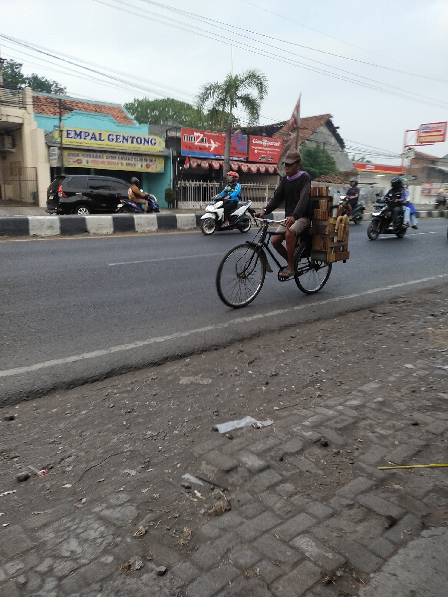 In Kemlakagede, Indonesia, a man rides a bicycle laden with wooden crates filled with bamboo poles along a busy street. The man, wearing a brown cap and a dark long-sleeved shirt, pedals the bicycle with his right leg extended. He is wearing shorts and sandals. The bicycle has a wooden frame extending from the rear to hold the cargo. The road is asphalt and appears to be in moderate condition, with some cracks and debris visible on the side. Several motorcycles are also present on the road, suggesting active traffic. To the left of the road, buildings with signs are visible. One building has a blue facade with yellow lettering that reads "EMPAL GENTONG." Another building displays a red and white sign for "Lion Parcel." The sky is overcast, with a few power lines crisscrossing overhead. The scene depicts a typical day in a bustling Indonesian city.