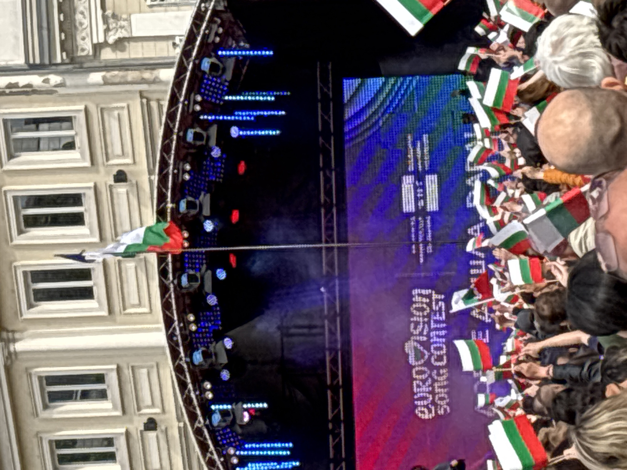 A crowd holds Bulgarian flags in front of a stage during the Eurovision Song Contest in Sofia, Bulgaria. The stage has the words "Eurovision Song Contest" on the screen. Blue spotlights are on the left side of the stage. The facade of a building is visible on the left.