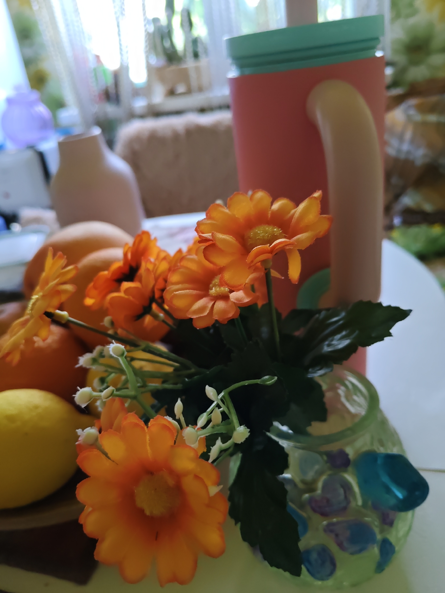 The image depicts a still life arrangement on a white surface, likely a table. In the foreground, there are several objects: a cluster of artificial orange flowers with green leaves and small white buds. One flower is particularly prominent, showcasing its orange petals and yellow center. To the left of the flowers, there are at least two round, yellow objects, appearing to be lemons. Behind the flowers and fruits, there are more round, orange objects, resembling oranges. A delicate, light green glass vase with colorful decorative stones (blue and purple) holds the flowers. In the background, a tall, pink cylindrical object with a light yellow handle and a mint green lid stands upright. To the left of this, a light beige vase with a narrow neck is visible, and further left, a translucent purple object. The overall environment appears to be indoors, with soft natural light filtering in from a window, creating a bright and airy atmosphere. The background is blurred, suggesting a shallow depth of field, but hints of curtains, furniture, and possibly plants can be discerned. There is no indication of people or any actions taking place. The scene does not provide any specific clues to its location within the city of Rostov-on-Don, Russia.