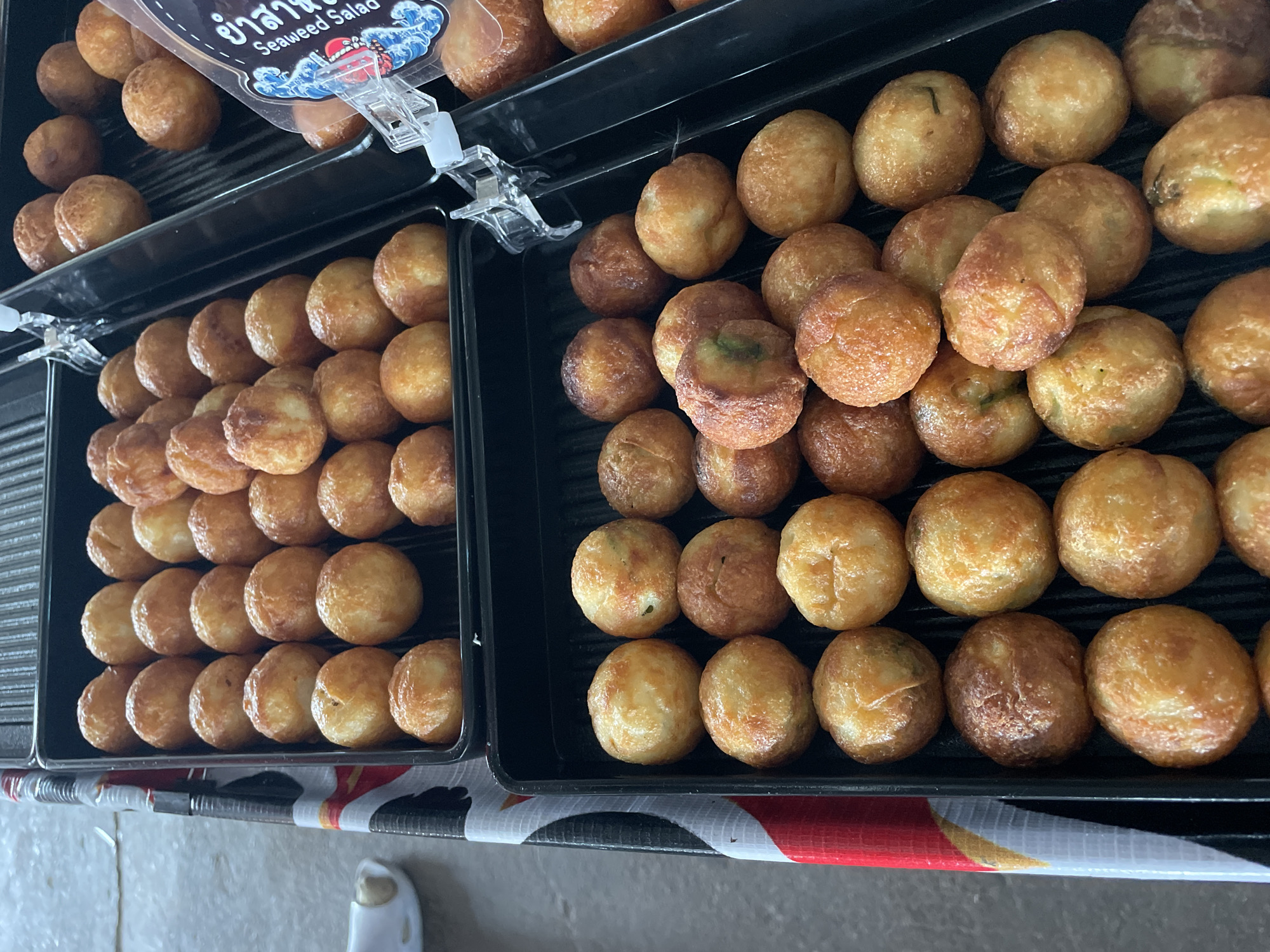 The image shows a close-up view of two black trays filled with golden-brown, round fried snacks, likely takoyaki. The snacks are arranged in rows within the trays. The top tray contains more snacks than the bottom tray. A clear plastic packet with Thai writing and the words "SEAWEED SALAD" is visible on the right side of the top tray. The background appears to be a light-colored, possibly concrete surface, and a black and white patterned fabric is visible on the left side. The overall setting suggests a food stall or market environment, potentially in Nong Prue, Thailand. No people are visible in the image.