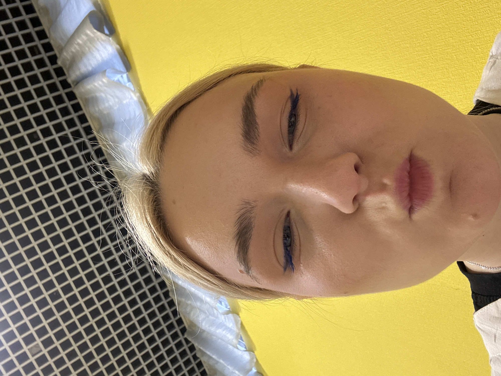A close-up, low-angle shot features the face of a young blonde woman against a bright yellow background. Her head is tilted, and she is looking down and to the right. Her eyes are made up with dark eyebrows and blue eyeliner. Her lips are slightly parted, and she appears to have a subtle smile. To the left of the woman, a black and white grid pattern is visible, with some light-colored, soft material draped over it, appearing to be textured or folded. This suggests an indoor setting with some distinct visual elements in the background. The lighting is bright, illuminating her face and the yellow background. No text is visible in the image. The overall impression is one of a casual, personal photograph.