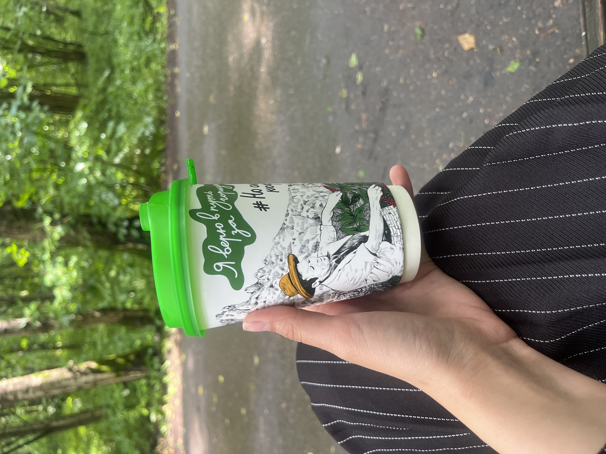 A person's hand holds a disposable coffee cup with a green lid. The cup is decorated with a drawing of a man in a hat and a landscape, as well as text in Russian. The person is wearing a black and white striped garment. The background is a blurry park setting with trees and a paved path. The image appears to be taken in a park in Moscow, Russia.