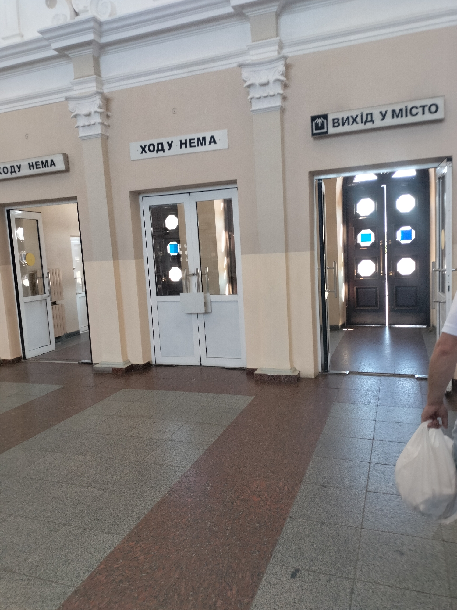 The image depicts an interior hallway with tiled flooring, likely at a transportation hub in Chernihiv, Ukraine. Two doorways are visible. The doorway on the left is partially open, revealing a white door and a circular yellow sign. Above this doorway, a sign reads "ХОДУ НЕМА" (Khodu nema), which translates to "No way" or "No passage." To the right of this, a second doorway is also visible with a similar sign above it, also reading "ХОДУ НЕМА." Further to the right, a larger, darker double door is present, with a sign above it that reads "ВИХІД У МІСТО" (Vykhid u misto), meaning "Exit to the city." This exit door has octagonal glass panes that are illuminated from behind and two blue square markings. A person, partially visible on the right side of the frame, is holding a white plastic bag. The hallway walls are painted a light, peachy color and feature decorative architectural elements along the top. The floor is tiled with a pattern of dark red and grey granite-like tiles. Sunlight is streaming through the exit door, casting shadows on the floor.