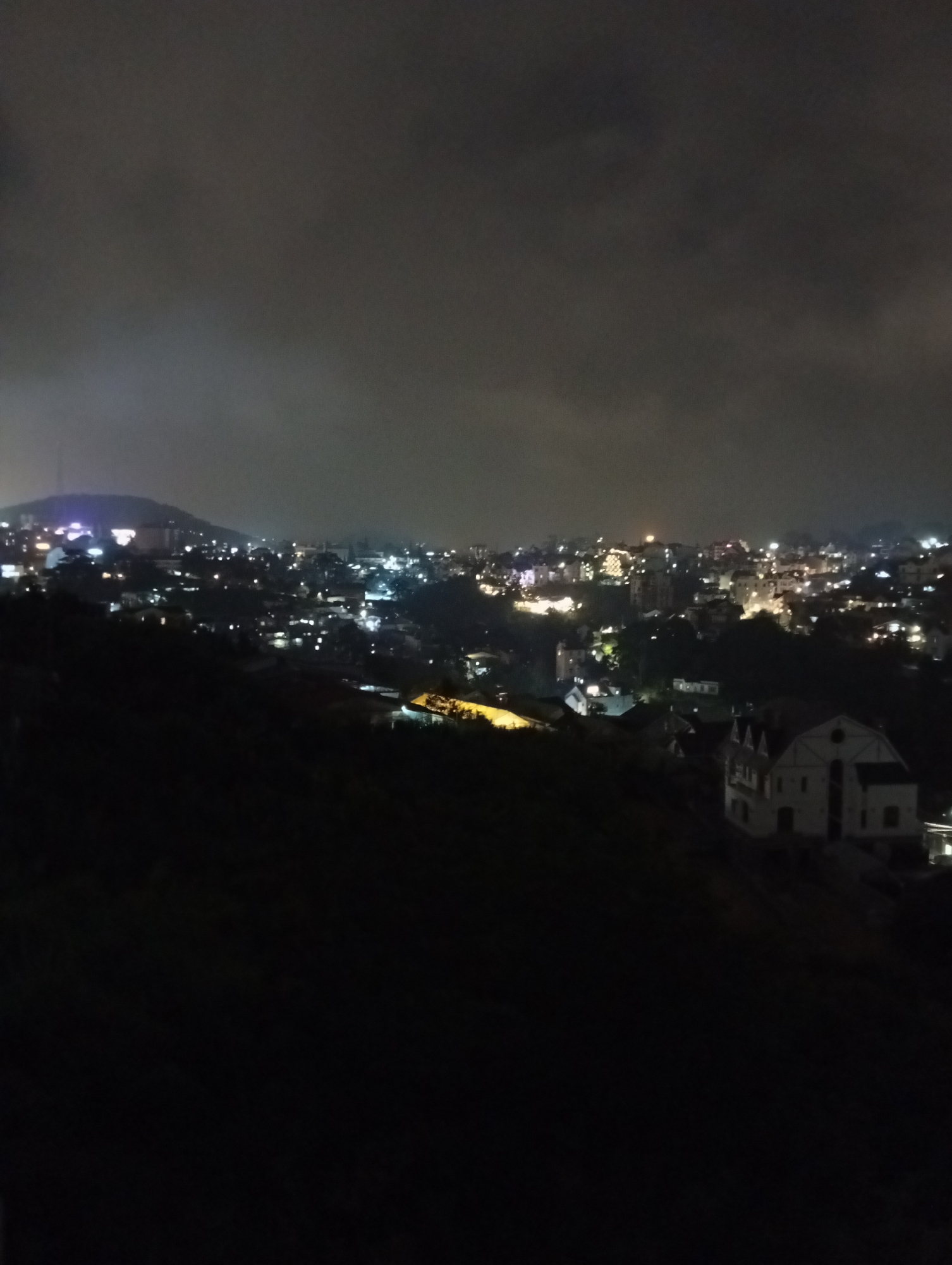 This is a nighttime, high-angle shot of a hillside city illuminated by artificial lights under a cloudy sky. The foreground is dominated by the dark silhouette of a steep slope, obscuring much of the immediate surroundings. In the mid-ground, several buildings are visible, including a prominent, light-colored structure with multiple gables, reminiscent of European architecture. This building appears to be a house or a small hotel, with its windows softly lit.
Beyond this, the city sprawls across rolling hills, a dense collection of buildings marked by numerous glowing windows and streetlights. The overall impression is of a vibrant urban landscape set against a dark, moody sky. The distant hills are faintly outlined, with a scattering of lights indicating further development. A brighter cluster of lights on a distant hilltop suggests a significant landmark or a more densely populated area. The time of day is clearly night, and the clouds overhead create a soft, diffused illumination, possibly indicating overcast conditions or fog. There are no visible people or text in the image. The scene conveys a sense of quiet urban life unfolding after dark, with the lights acting as beacons in the night. Given the location context of Da Lat, Vietnam, the architecture of the prominent building in the mid-ground could suggest areas with historical European influences, common in hill stations.