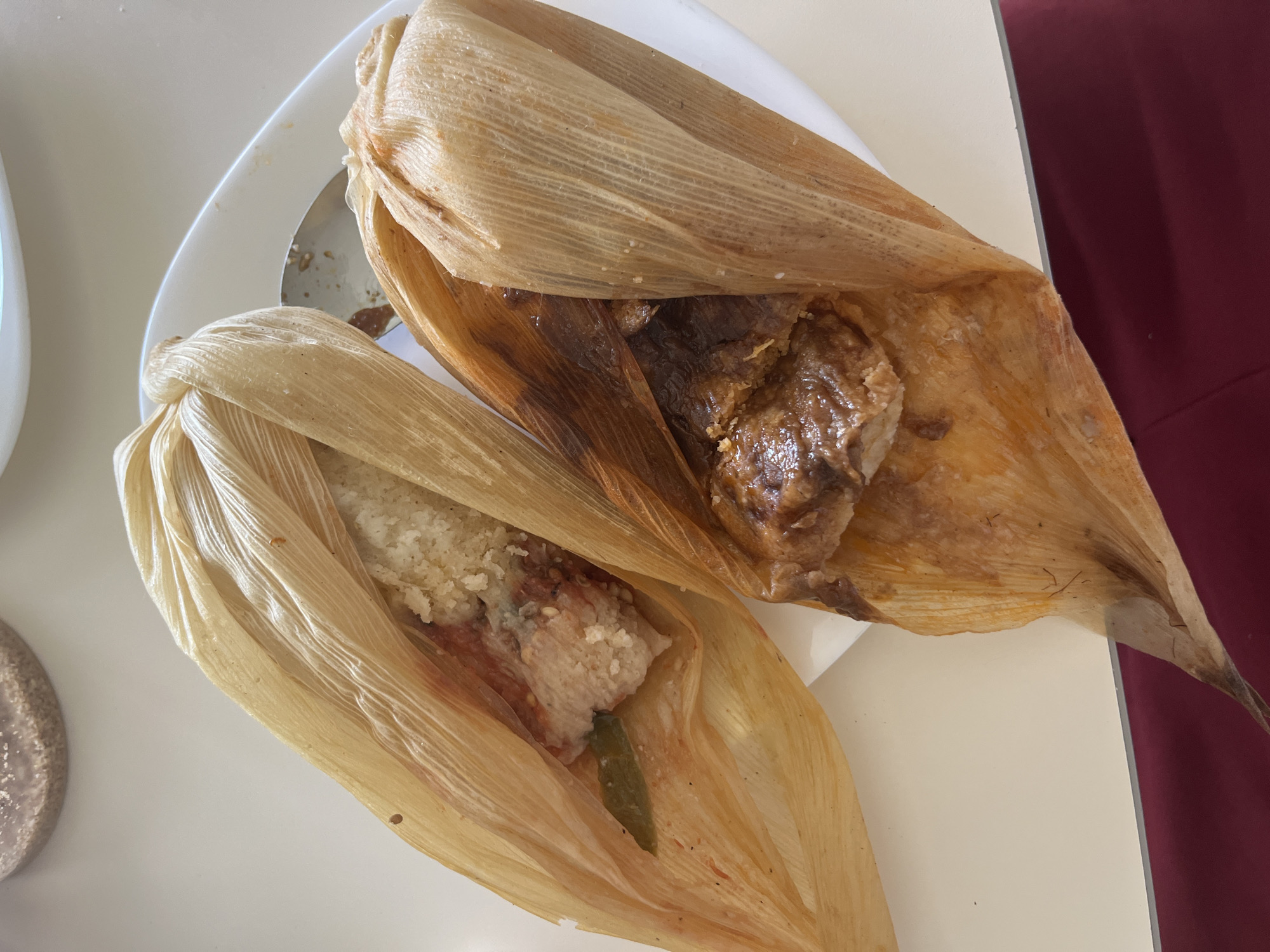 The image shows two tamales on a white plate. One tamale contains a dark, saucy meat filling. The other tamale appears to have a lighter-colored filling with what looks like a green pepper. The tamales are wrapped in dried corn husks. The plate sits on a white surface, with a maroon-colored object partially visible in the background.