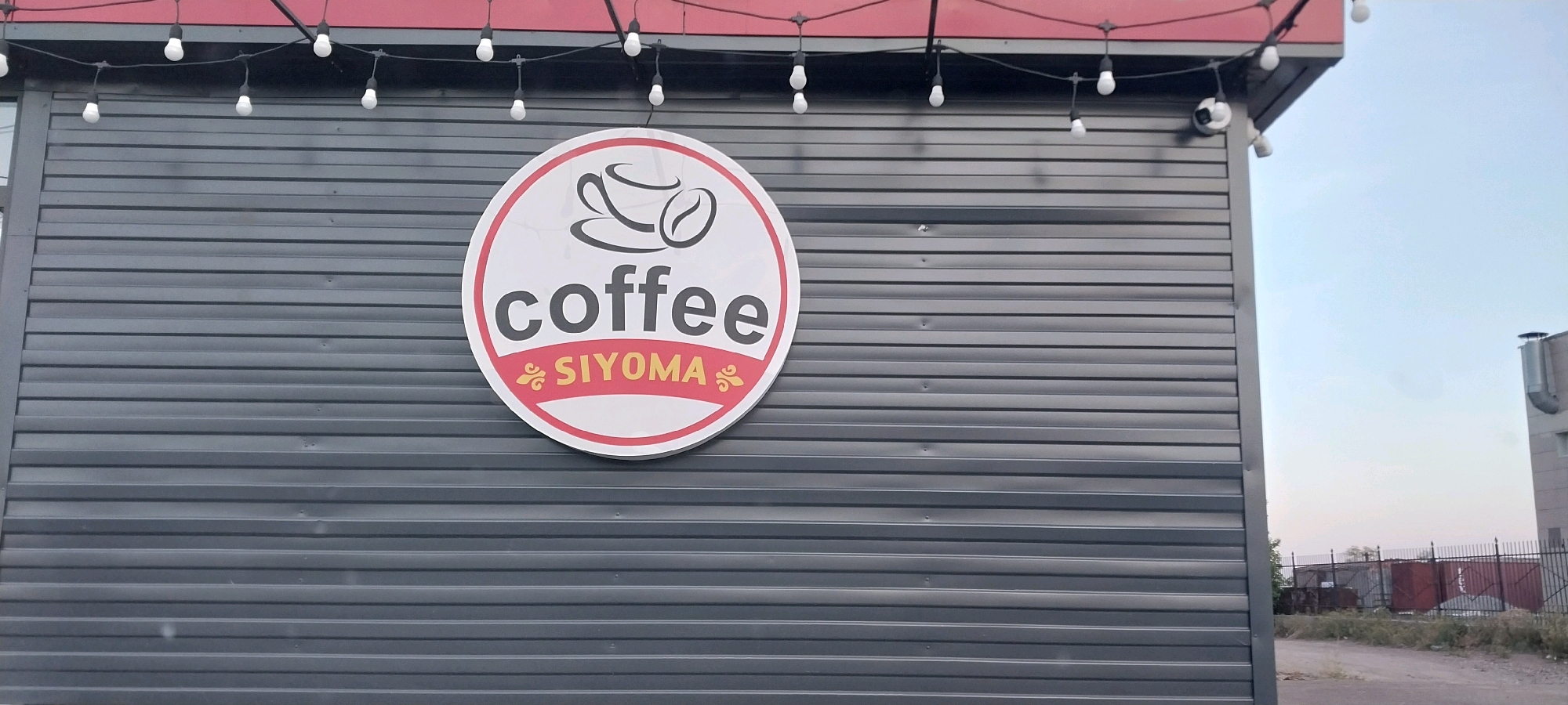 The image shows a close-up of a coffee shop sign on the exterior wall of a building. The sign is circular with a red border and a white interior. It features a stylized image of a coffee cup and a coffee bean at the top. Below the image, the word "coffee" is written in large, bold black letters. Underneath "coffee," the word "SIYOMA" is written in smaller, yellow letters with decorative red accents on either side. The building wall is made of dark gray, corrugated metal. A string of white, bare light bulbs is visible above the sign, attached to a red overhang. A security camera is mounted on the upper right corner of the building. In the background, a clear sky can be seen, along with some industrial structures and a fence.