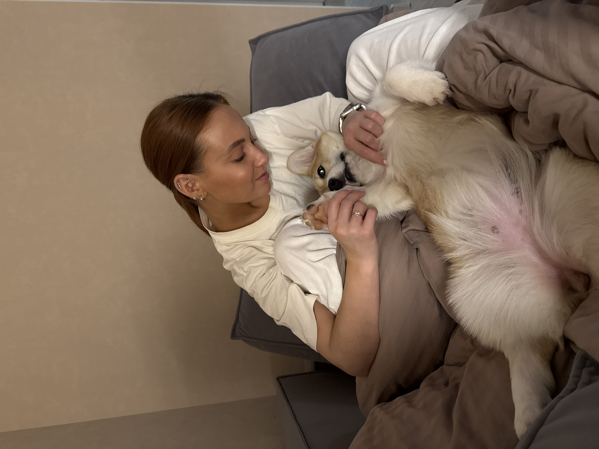 A woman lies on a couch, holding a dog upside down on her chest. The woman has auburn hair pulled back in a ponytail, fair skin, and multiple piercings in her left ear. She wears a long-sleeved white shirt, a watch on her left wrist, and a ring on her left ring finger. She is looking at the dog with a gentle expression. The dog, a corgi, is on its back with its belly exposed. The dog has white and tan fur. The couch is covered in grey and brown bedding. The background is a plain beige wall. The location is not discernible from the image.