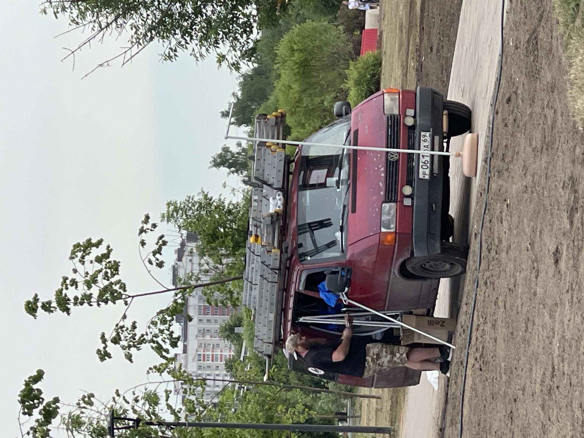 A man wearing a black t-shirt, camouflage shorts, and a baseball cap stands next to the open side door of a red Volkswagen van. The van has multiple ladders secured to its roof rack. The man is holding what appears to be an electrical conduit or pole. Inside the van, blue tarpaulin is visible. A cardboard box labeled "Zelli" rests on the ground near the van's door. The vehicle's license plate, visible at the front, reads "P 061 JA 69". The scene takes place on a paved pathway surrounded by dirt and grass. In the background, there are trees, a tall building with multiple windows, and a lamppost. The sky is overcast. The overall setting suggests an outdoor work or construction site in a park or urban area, likely in Moscow, Russia, given the vehicle's license plate format.