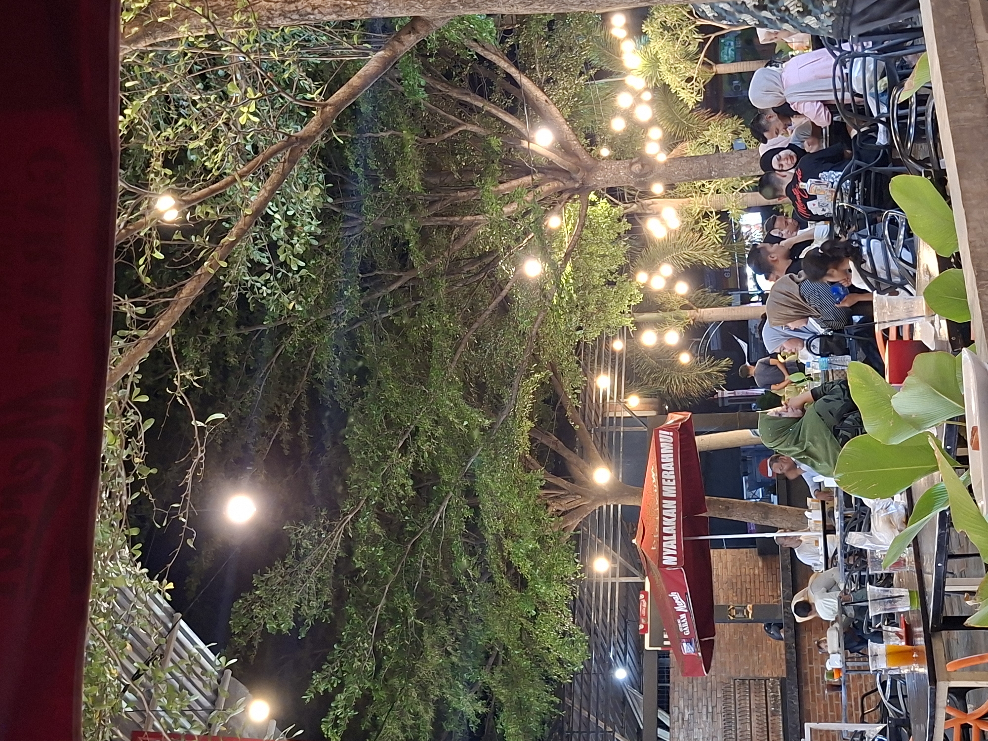 The image captures an outdoor scene, likely a restaurant or café, in Lhokseumawe, Indonesia. Several people are seated at tables, some engaged in conversation, while others seem to be enjoying their meals or drinks. The setting is partially shaded by large trees adorned with strings of lights, creating a warm and inviting atmosphere. An umbrella bearing Indonesian text is visible, suggesting a local establishment. The architecture in the background hints at an urban environment.