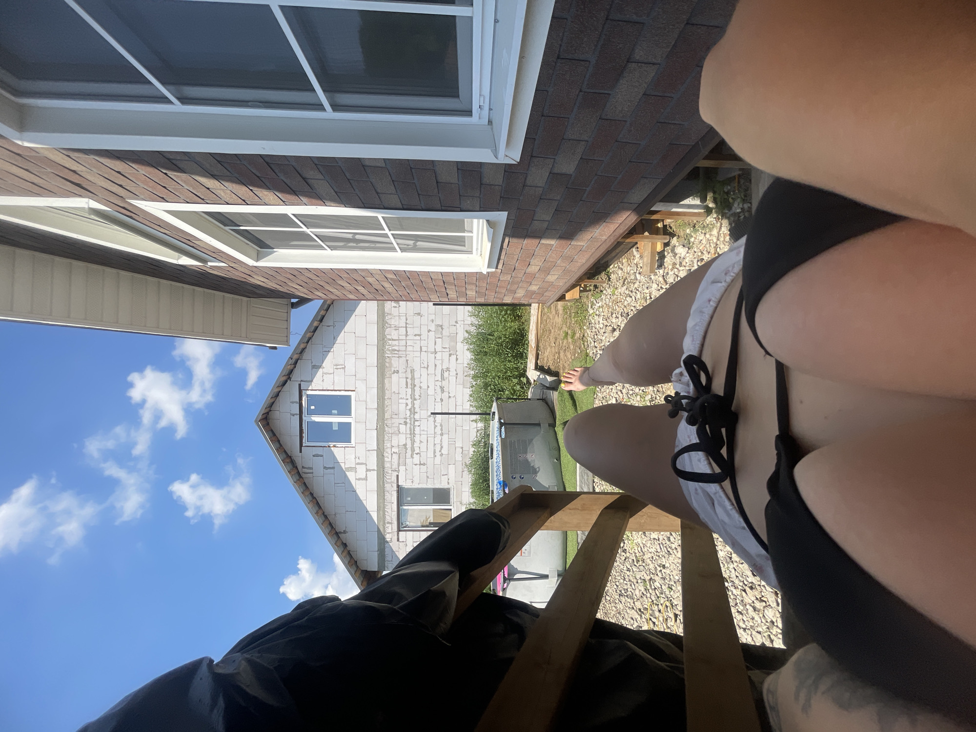 The image shows a person lying on a wooden deck in a backyard. The person is wearing a black bikini and white shorts. In the background, there is a partially constructed house made of white brick, and a finished house with dark brown brick siding. An inflatable swimming pool is visible between the two houses. The ground is covered with gravel and grass. The sky is blue with some white clouds. The person's legs and torso are the main focus of the image. The location is identified as Mosmetalkonstruktsiya Ont, Russia.