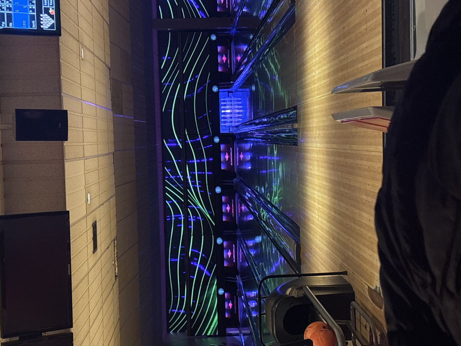 The image, originally rotated 90 degrees clockwise, captures an indoor bowling alley, likely located in London, United Kingdom. The scene depicts a "cosmic bowling" or "glow bowling" session, characterized by a dark environment illuminated with vibrant neon and UV lights, suggesting an evening or nighttime activity.
On the left, a light-colored, brick-patterned wall features several mounted screens. The topmost screen displays a bowling scoring system with visible text including "SCORE," "LANE," player names "LENZ" and "JACK," and numerical scores like "6-1" and "6-6," along with a graphic of bowling pins. Below it, a dark, rectangular monitor is also affixed to the wall.
The central and dominant part of the image shows multiple bowling lanes brightly lit with glowing green wavy lines and purple/blue accents. These neon patterns extend along the entire length of the lanes, creating a dynamic visual effect. At the far end of the lanes, sets of bowling pins are visible on their respective pin decks, also highlighted by the colorful lighting.
To the right, the light-toned wooden bowling lane surface is prominent, leading towards the player area. A person, dressed in dark attire, is partially visible on the far right edge of the frame, suggesting their presence in the playing zone. In the lower right foreground, an orange bowling ball rests in a metal ball return rack, indicating active play. The overall atmosphere is lively and modern.