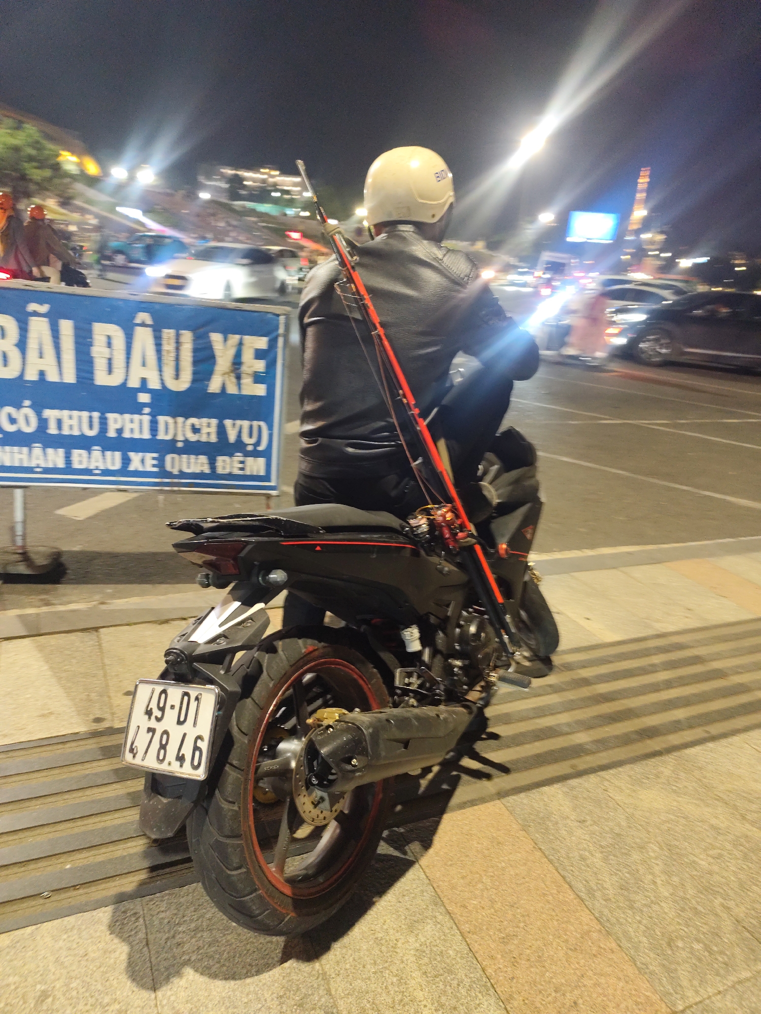 In Da Lat, Vietnam, at night, a person wearing a white helmet and a black leather jacket sits on a dark-colored motorcycle. The motorcycle has a red accent on its seat and a fishing rod secured vertically to its side. A license plate reading "49-01 478.46" is visible on the rear of the motorcycle. In the background, there are blurred lights from streetlights and buildings, indicating an urban environment. A blue sign with white Vietnamese text is partially visible to the left of the motorcycle, reading "BÃI ĐẬU XE" which translates to "PARKING LOT." The ground is paved with stone tiles and there are horizontal metal strips on the ground in the foreground.