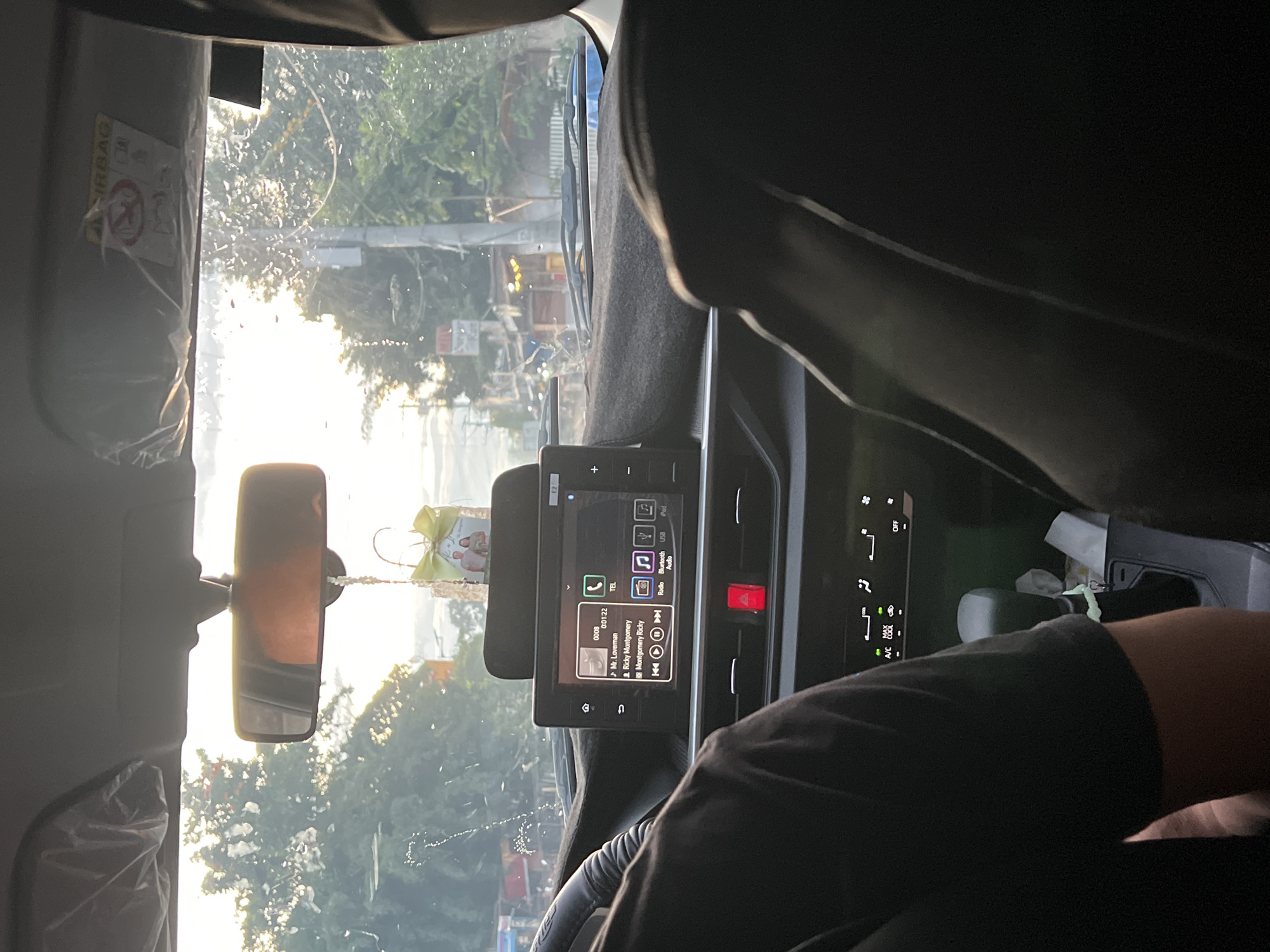 The image shows a view from inside a car, presumably the driver's perspective, looking out through the windshield. Outside, there is a street scene with trees and buildings, possibly near a body of water. The sun is shining brightly, creating glare on the windshield.
Inside the car, the dashboard and infotainment system are visible. The screen displays what appears to be a music player interface. There is a rear-view mirror. The driver, partially visible, is wearing a dark t-shirt. Other car components are visible such as airbag label, air conditioning vent controls.
The location is identified as Davao City, Philippines.
