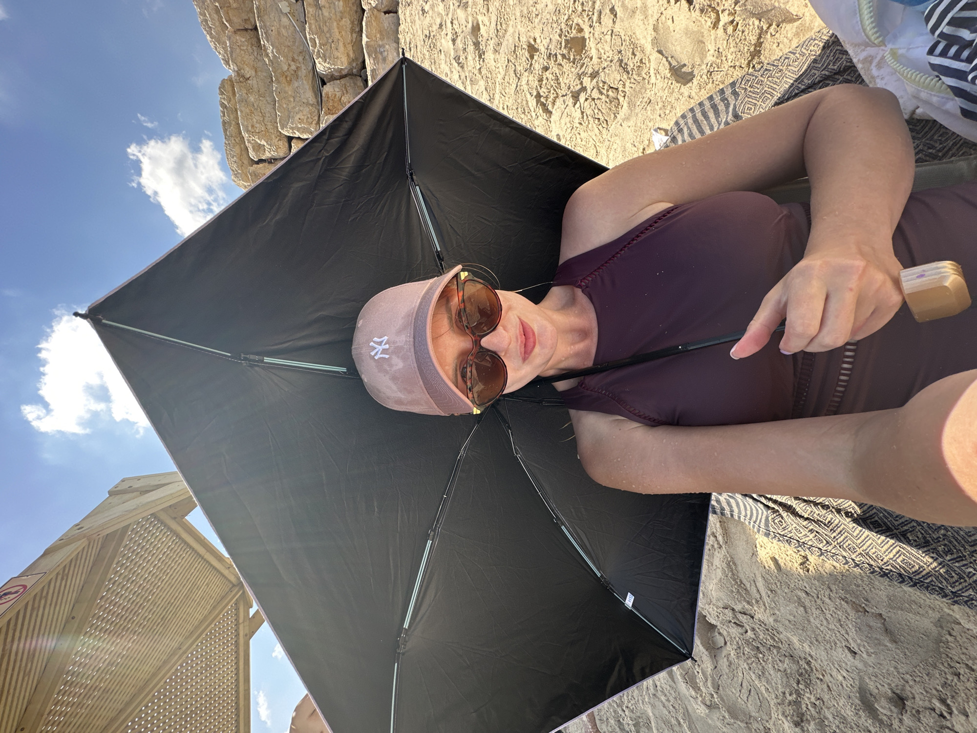 A woman is sitting on a sandy beach in Tel Aviv, Israel, under a black umbrella. She is wearing a maroon swimsuit and a pink baseball cap. She is also wearing sunglasses. The sun is shining and there are a few clouds in the sky. There is a wooden structure with a lattice roof to her left, and a stone wall behind her. She is holding the handle of the umbrella with her right hand. There is a towel with a geometric pattern on her lap. The overall scene is one of a relaxing day at the beach.