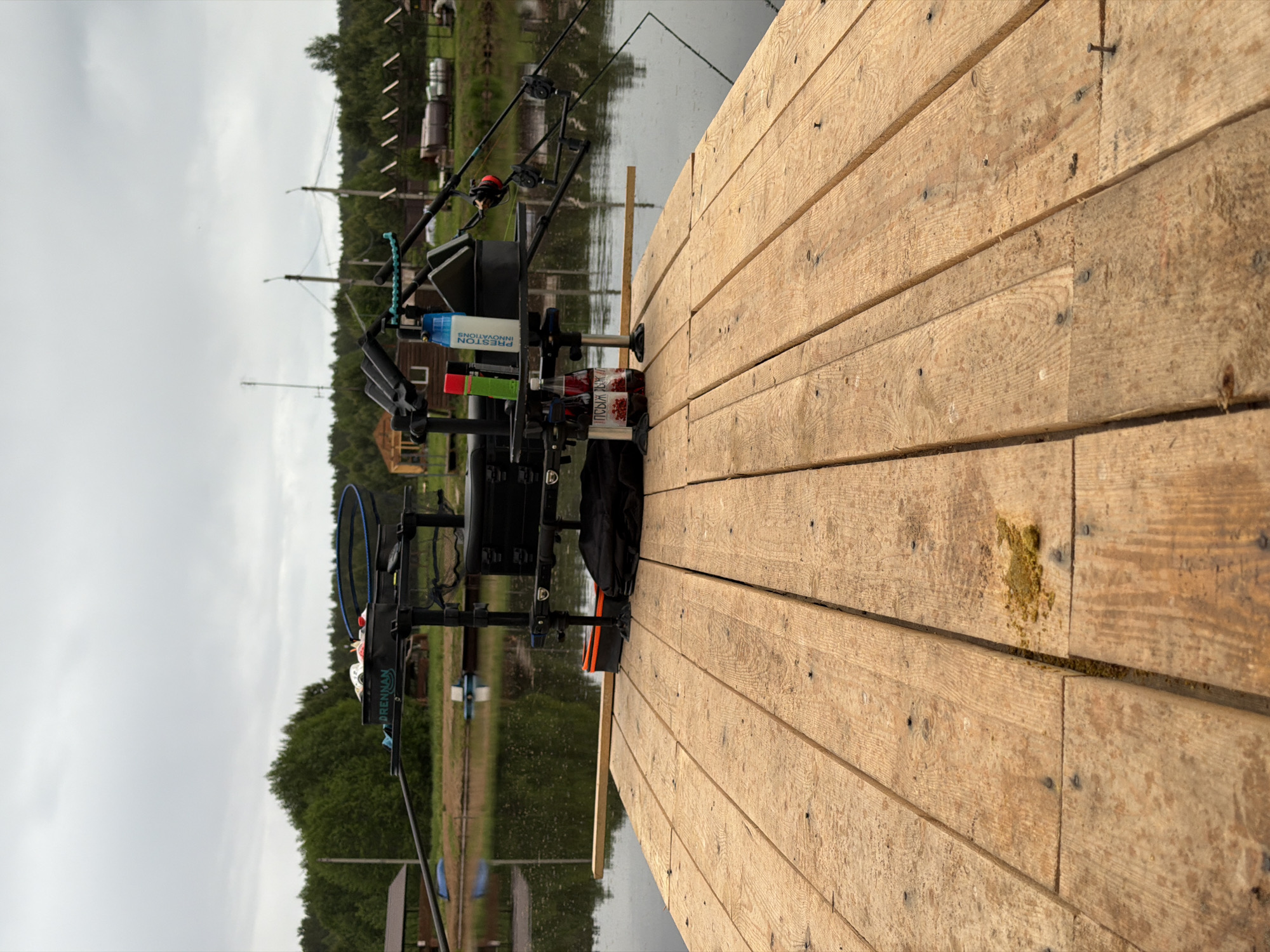 The image depicts a fishing setup on a wooden pier or platform. The fishing equipment includes rods, a seat box with "Drennan" visible on the side, and various accessories like a bait container labeled "Preston Innovations". A bottle of juice with a Cyrillic label and a black bag are also present. The location appears to be near a body of water, possibly a lake or river, with forested areas in the background and structures that could be small buildings or cabins. Based on the prompt, the location is Прикамский, Russia. The scene is presented from an off-angle perspective, focusing on the fishing equipment and the wooden pier.