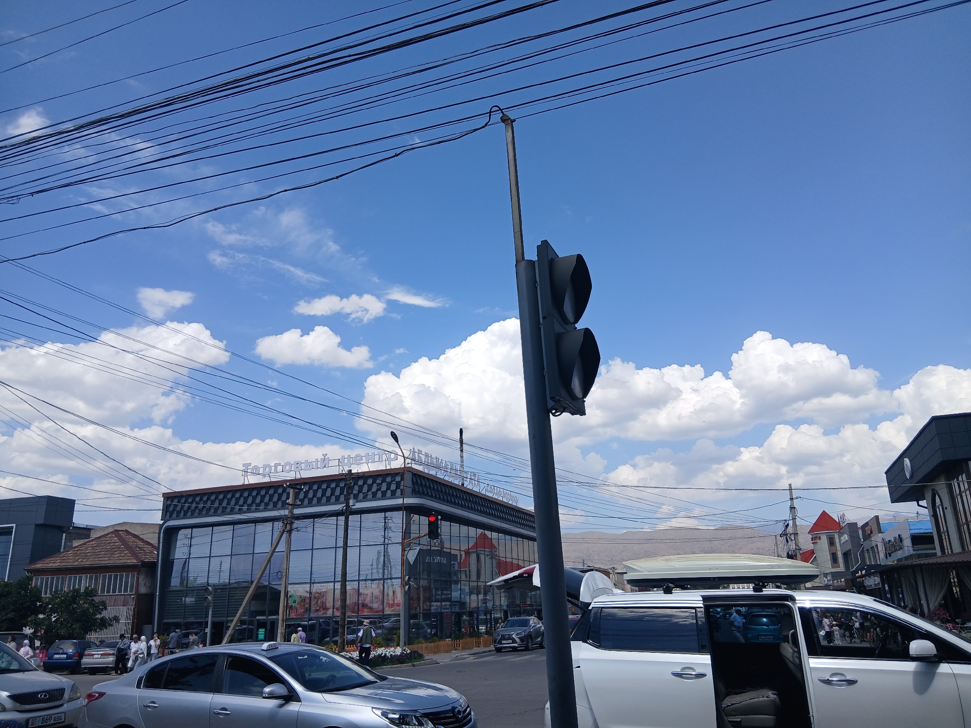 This is a street-level, eye-level, medium shot taken in Talas City, Kyrgyzstan. The image depicts a street scene with several vehicles, buildings, and a traffic light. In the foreground, there are two cars; a silver sedan on the left and a white van with its doors open on the right. The white van has a roof-mounted cargo carrier. Behind the vehicles, a building with a checkered facade and large glass windows is visible. This building has signage in Cyrillic script, which reads "Торговый Центр" (Trading Center). A traffic light with a red light illuminated is positioned on a pole in the mid-ground. Several people are walking on the sidewalk in front of the building. The sky is blue with scattered white clouds, and numerous power lines are strung across the upper portion of the frame. In the background, there are more buildings and hills. The overall impression is a bustling urban environment on a clear day.