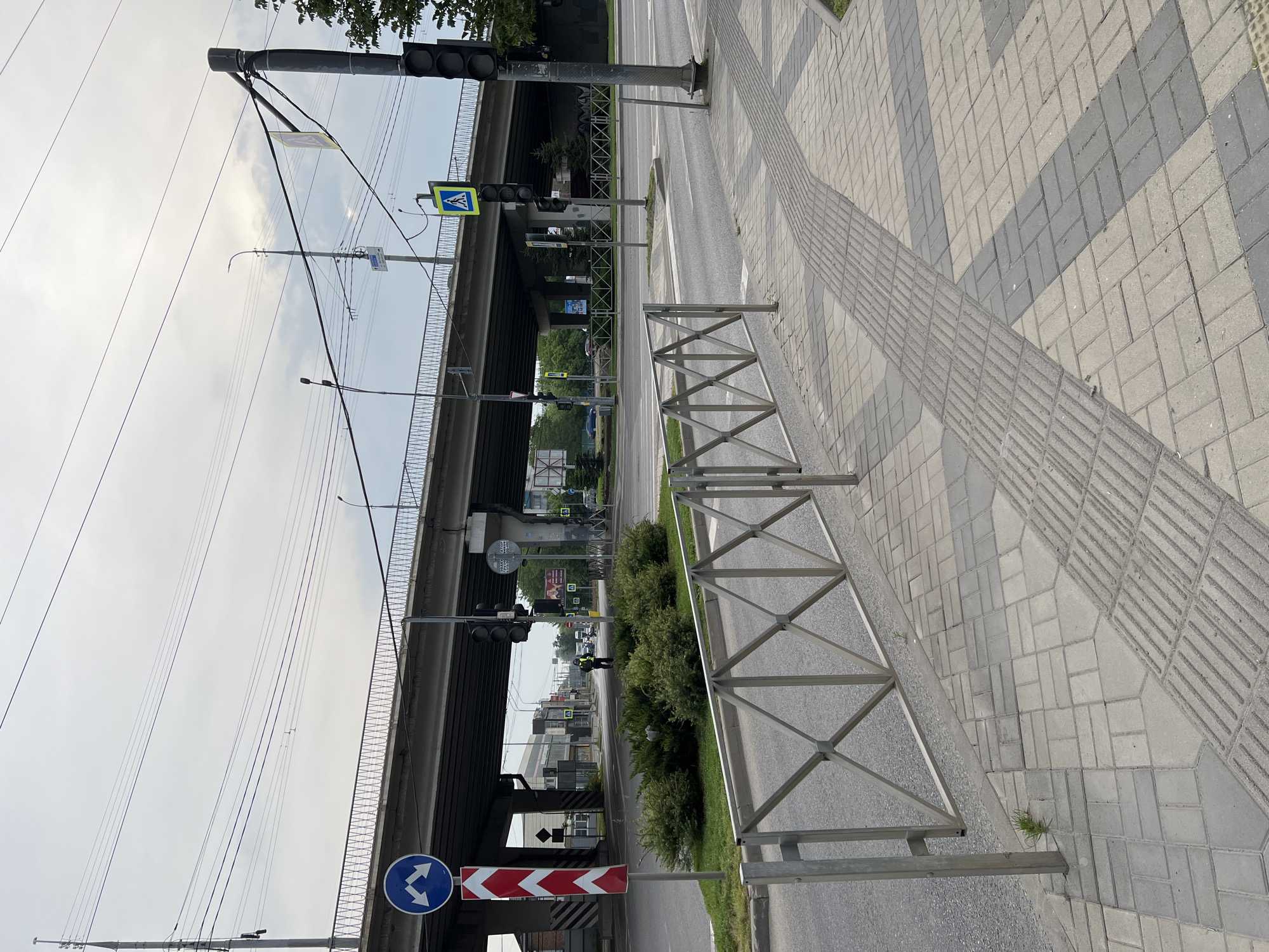 The image shows a street scene in Krasnodar, Russia. A gray metal fence separates the sidewalk from the road. The sidewalk is paved with gray concrete blocks and has a tactile paving strip. An overpass is visible in the background. Traffic lights and road signs are present. A bush grows along the roadside behind the fence. The sky is overcast.
