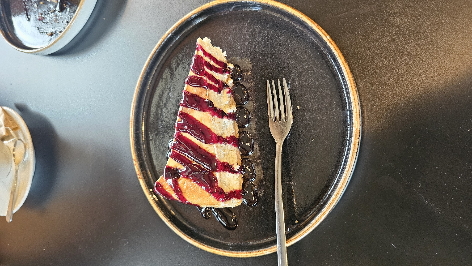 The image shows a slice of cake topped with a dark berry sauce, presented on a black plate with a bronze rim, alongside a fork. The cake slice is triangular in shape. The sauce appears to be drizzled across the top and pooling around the cake. The photograph was taken indoors, likely in a restaurant. The table surface is black. A portion of another plate and silverware are visible in the background. The location is Varna, Bulgaria.