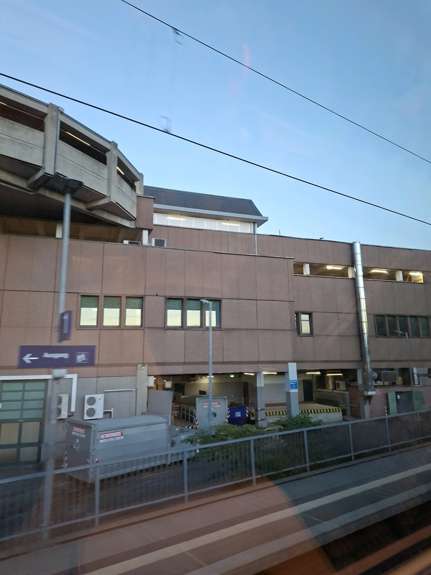 The image shows a multi-story parking garage in Koblenz, Germany, likely near a train station given the overhead wires. The exterior of the building is clad in brown panels, with horizontal bands of windows and ventilation grates. On the lower level, a large grey dumpster is visible next to a doorway with a green glass panel. A purple sign with a white arrow pointing left and the word "Ausgang" (exit) is mounted on a pole. The sky is a pale blue with some subtle pinkish hues, and power lines crisscross the upper portion of the frame. The scene is captured at dusk or dawn, with artificial lights visible inside the parking garage.