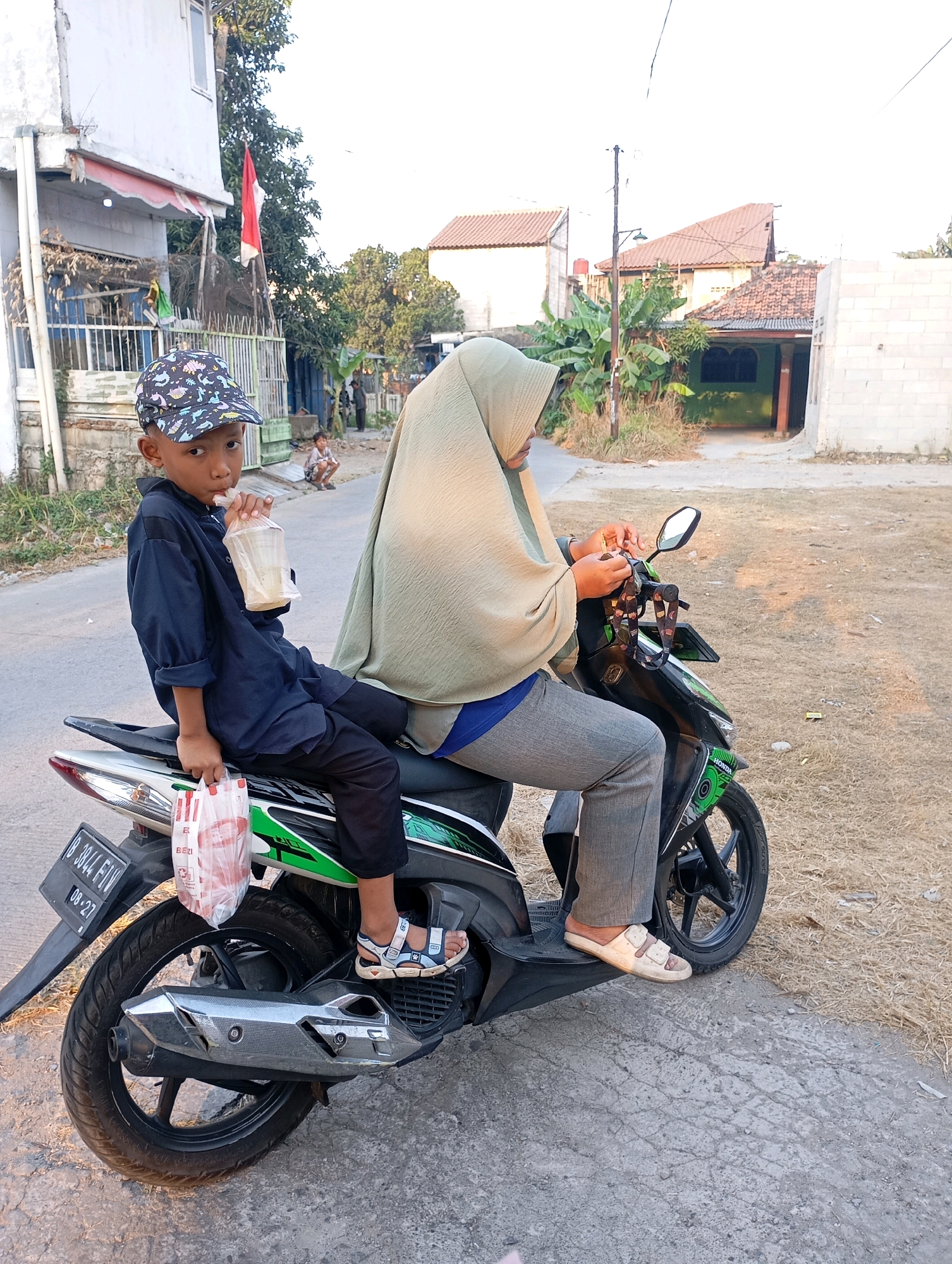 In Babelan, Indonesia, a young boy wearing a black long-sleeved shirt, black pants, and a patterned baseball cap sits on the back of a black and green scooter. He is holding a clear plastic cup with a straw to his mouth, drinking a light-colored beverage. In front of him, a person wearing a light green hijab and gray pants, presumably an adult, operates the scooter. The adult's hands are visible near the handlebars. A white plastic bag containing red items is attached to the side of the scooter, near the boy's feet. The scooter is parked on a paved road with dry grass and dirt on the right side. In the background, there are houses with tiled roofs, a white wall, trees, and a red and white flag flying from a pole. A child can be seen sitting on the ground in the distance. The sky is overcast and light.