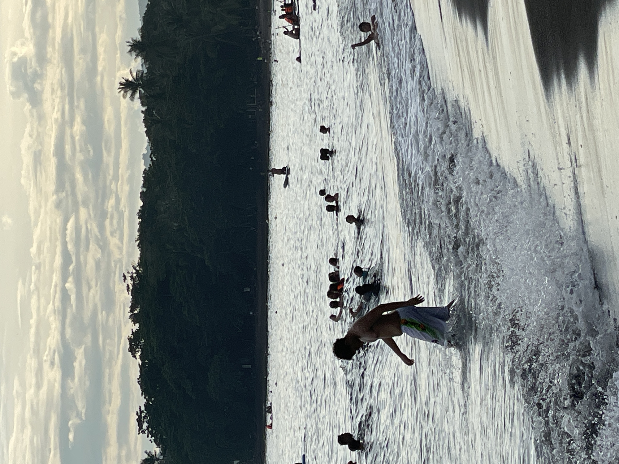In Dauin, Philippines, a group of people are enjoying the ocean near the shore. The foreground shows a man wading in the shallow water with waves breaking around him, his lower body submerged. He is wearing light-colored shorts with a patterned design. Behind him, a crowd of people are swimming and playing in the water, with some appearing to be in groups. Further out in the sea, there are individuals on paddleboards and a small boat with people. The background features a dense line of trees, likely a tropical forest, bordering the coastline. Above the trees, the sky is overcast with scattered clouds. The overall scene depicts a lively beach day with people engaged in various water activities.