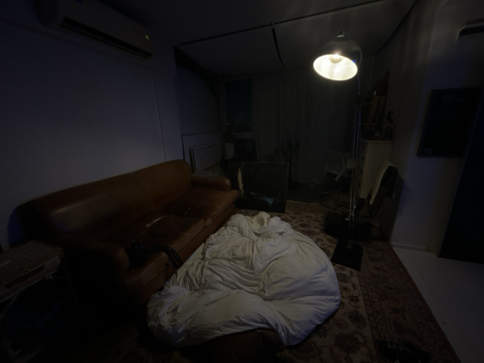 A dimly lit interior showcases a worn brown leather couch, partially obscured by a large, white beanbag. In the background, a standing lamp casts light on cluttered shelves and what appears to be art supplies. The room also features a dark painting, a chalkboard, and a glimpse of a window revealing the night outside. The floor is covered with a patterned rug, adding texture to the low-light setting.