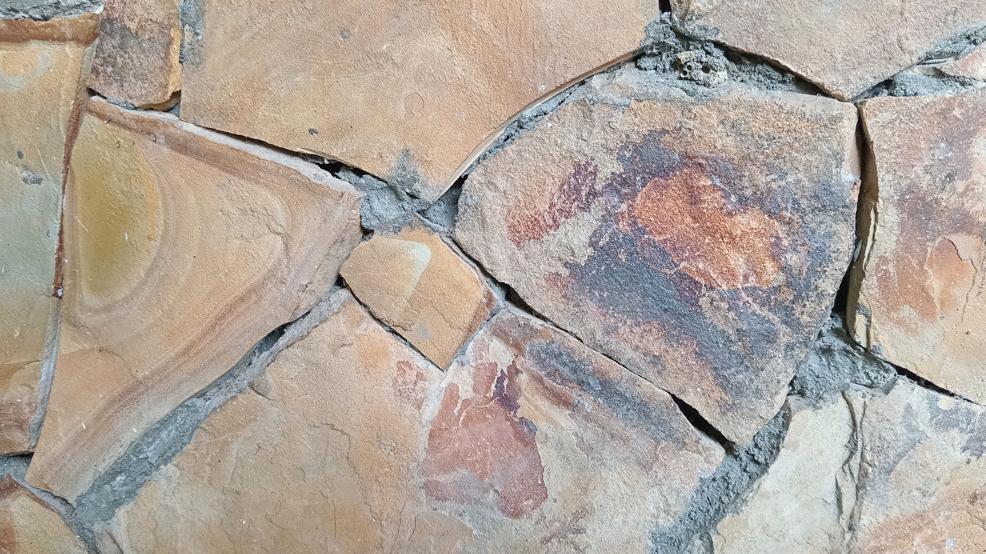 The image shows a close-up view of a stone wall. The stones are irregular in shape and size, with varying shades of brown, tan, and some reddish-orange and gray markings. They are mortared together with gray cement. The texture of the stones appears rough and natural. The scene depicts an exterior wall, likely part of a building or landscaping feature. There are no people or animals visible in the image. The lighting suggests it is daytime, with shadows indicating the direction of the light source. The location is specified as Randuagung, Indonesia.
