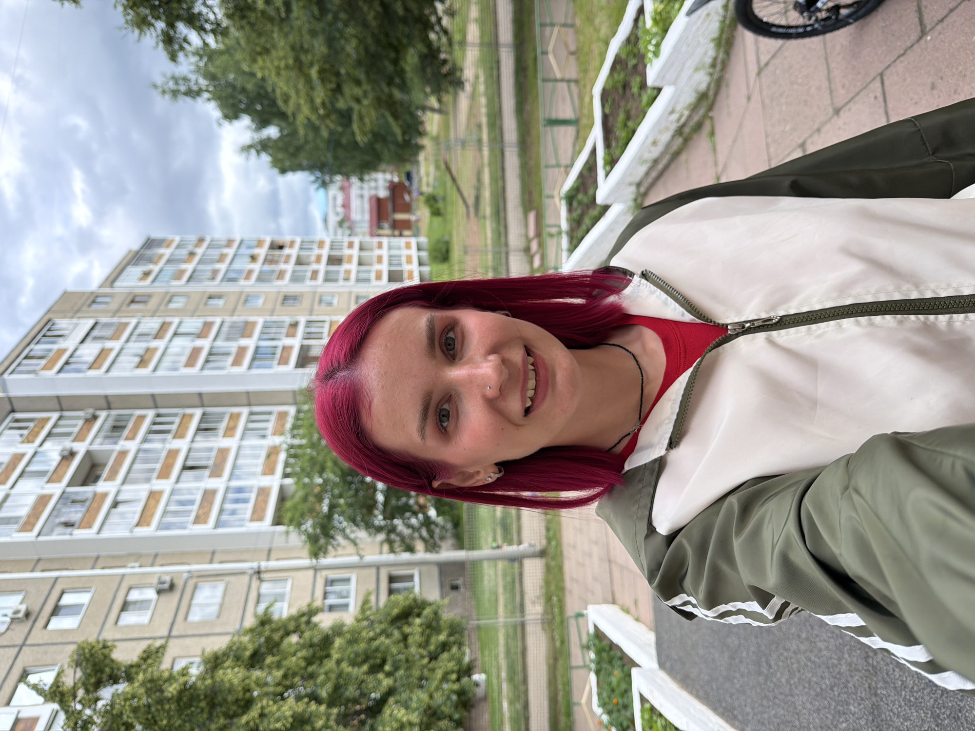 A young woman with vibrant pink hair smiles for a selfie. She is wearing a red shirt and a two-toned jacket, primarily white with green accents, and a black necklace. Her left ear is adorned with a silver hoop earring. She has a nose piercing.
The background reveals a slightly overcast sky and a large, multi-story apartment building with numerous windows. Trees with green foliage are visible in front of and around the building, and raised garden beds line a walkway. The ground appears to be a paved surface. The image is taken outdoors, likely during the daytime. The overall setting suggests an urban or suburban residential area.