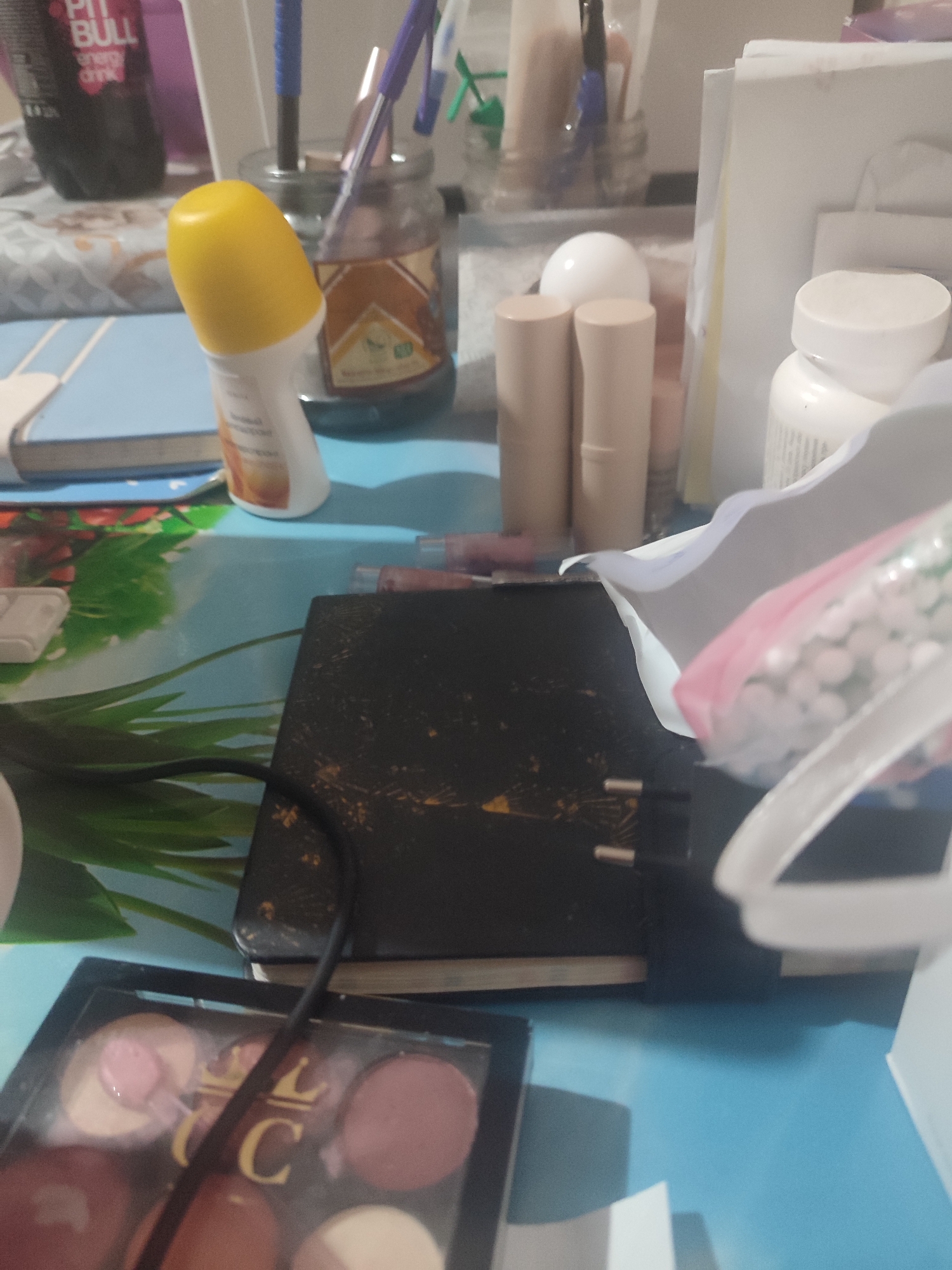 The image depicts a cluttered table surface with various items. In the foreground, a makeup palette with several circular pans of product, some with applicator wands, is visible. To the left, a dark, rectangular object, possibly a book or a notebook, is partially covered by a black cord. Behind this, a yellow-capped roll-on deodorant is standing upright. Further back, several cylindrical objects, likely lipsticks or cosmetic sticks, are arranged in a row. A glass jar filled with pens and pencils stands behind them. A dark bottle with a "PIT BULL ENERGY DRINK" label is on the far left. In the background, white papers and a white bottle with a red label are present. The table surface has a light blue and green patterned design. The scene appears to be indoors, likely a personal space given the nature of the objects. There is no information to ascertain the location in Chernivtsi, Ukraine.