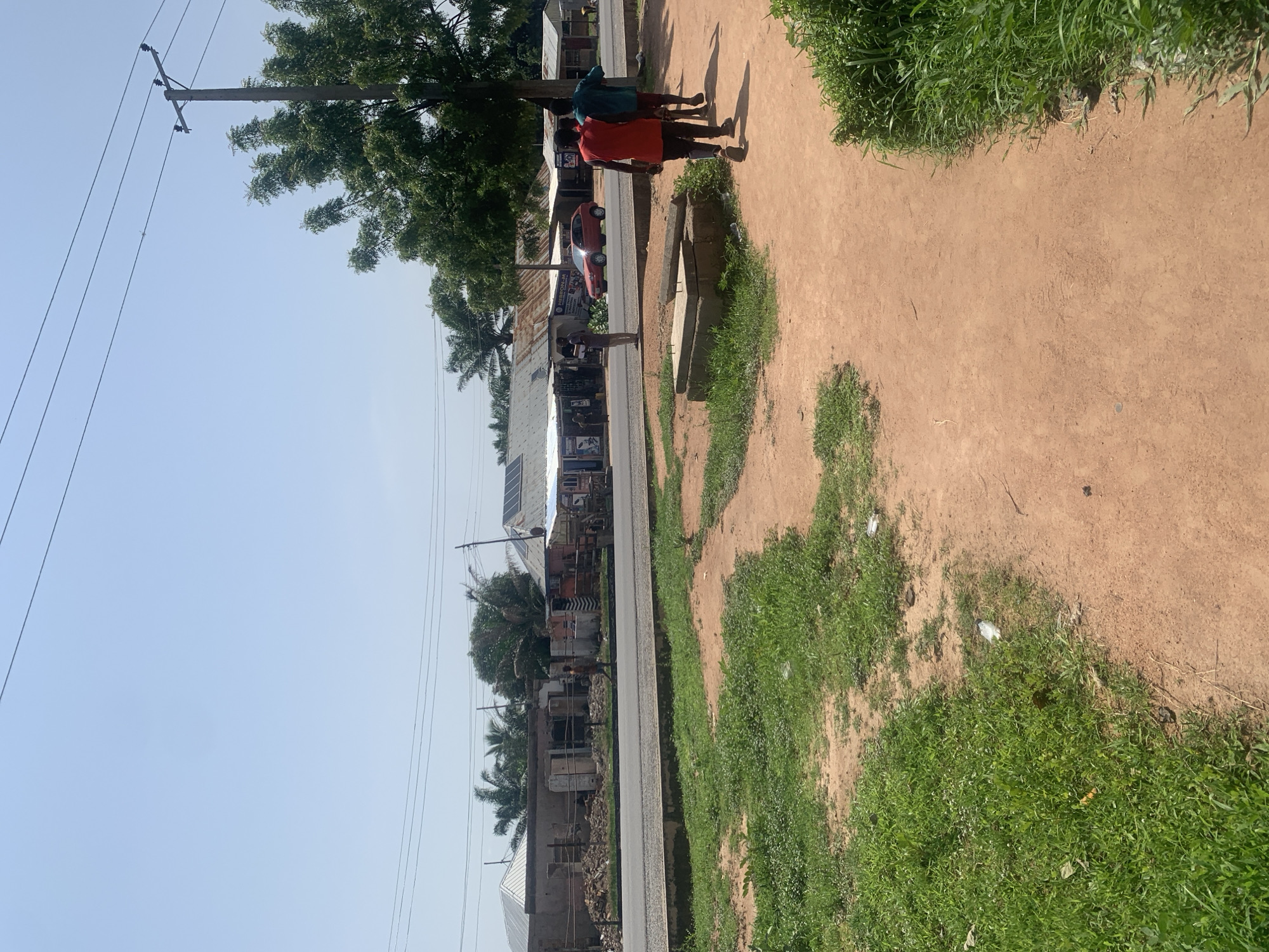 The image depicts a street scene in Jalingo, Nigeria. In the foreground, a dirt path runs alongside a patch of green grass. Further back, a paved road separates the foreground from a row of low-rise buildings. These buildings appear to be a mix of shops and residences, some with corrugated metal roofs and others with more modern, flat roofs, some equipped with solar panels. Debris, including bricks and what looks like building materials, is visible in front of some of the structures. Several people are present. Two individuals, dressed in red and blue shirts, are walking away from the camera on the dirt path. Another person, dressed in a red shirt, is standing near the road. A red car is parked on the road in front of one of the buildings. A utility pole with power lines stands on the right side of the frame, amidst lush green trees. The sky is clear and blue, indicating a sunny day.