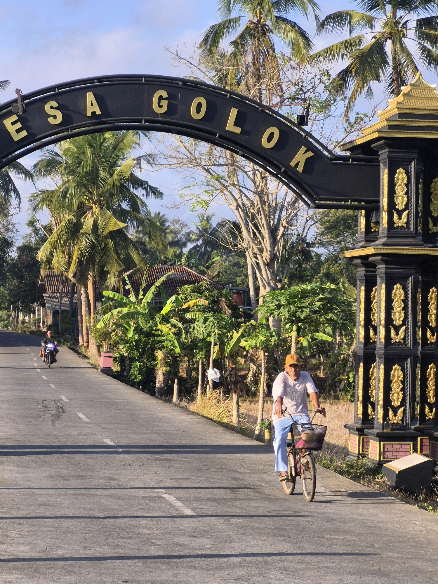 The image depicts an outdoor scene in Golok, Indonesia, featuring an ornate archway with gold lettering spelling "DESA GOLOK". To the right of the arch, a tall, black and gold decorative pillar stands. In the foreground, a man wearing a white t-shirt, blue pants, and an orange cap is riding a bicycle. The bicycle has a basket in front containing what appears to be a small object, possibly food. The man is on a paved road, which extends into the background. A motorcycle carrying a rider is visible further down the road on the left. The surroundings are lush with palm trees and other tropical vegetation, with hints of houses with tiled roofs visible behind the foliage. The sky is clear and blue, suggesting it is daytime. The overall impression is a rural or semi-rural setting in Golok, Indonesia, with elements of local architecture and daily life.