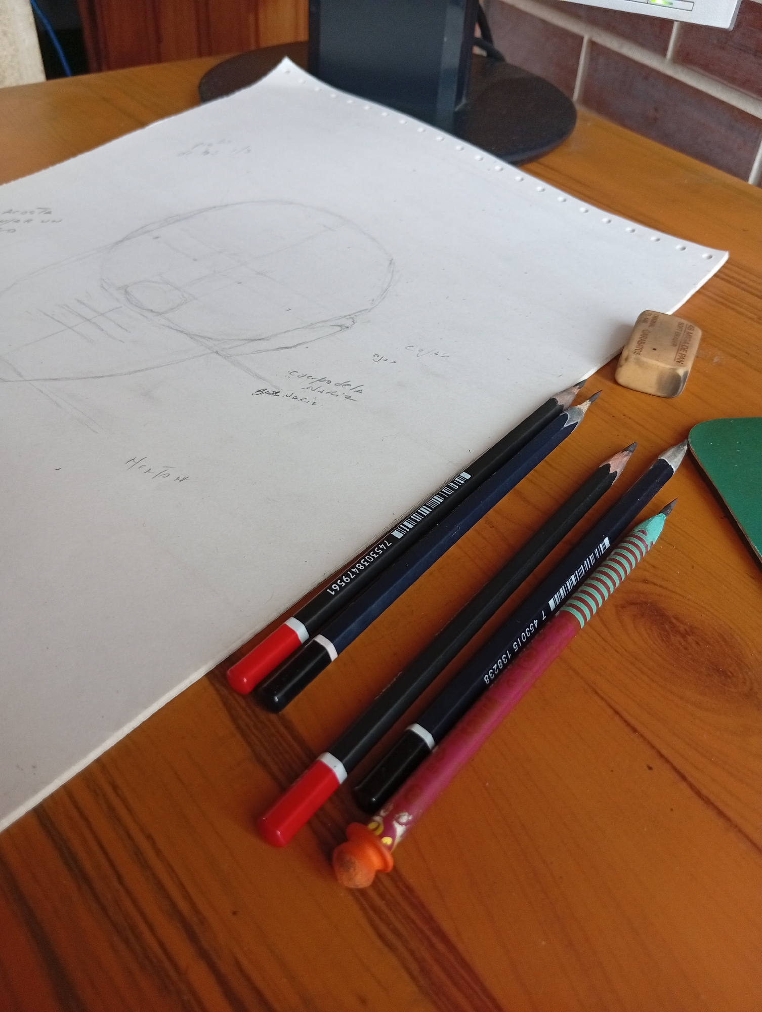 The image shows a wooden table with a white sheet of paper with a sketch on it, several pencils, and an eraser. The sketch appears to be a drawing of a head with some lines and annotations. The pencils are arranged in a row, with some having red caps. The eraser is a light brown color. In the background, a monitor and part of a brick wall are visible. The overall scene suggests an art or drafting workspace. The text on the eraser, "ACME DE PAN", is a brand of bread, suggesting this might be a repurposed eraser or an oddity in the workspace. The specific location in San Antonio de Los Altos, Venezuela cannot be determined from the visual information present in the image.