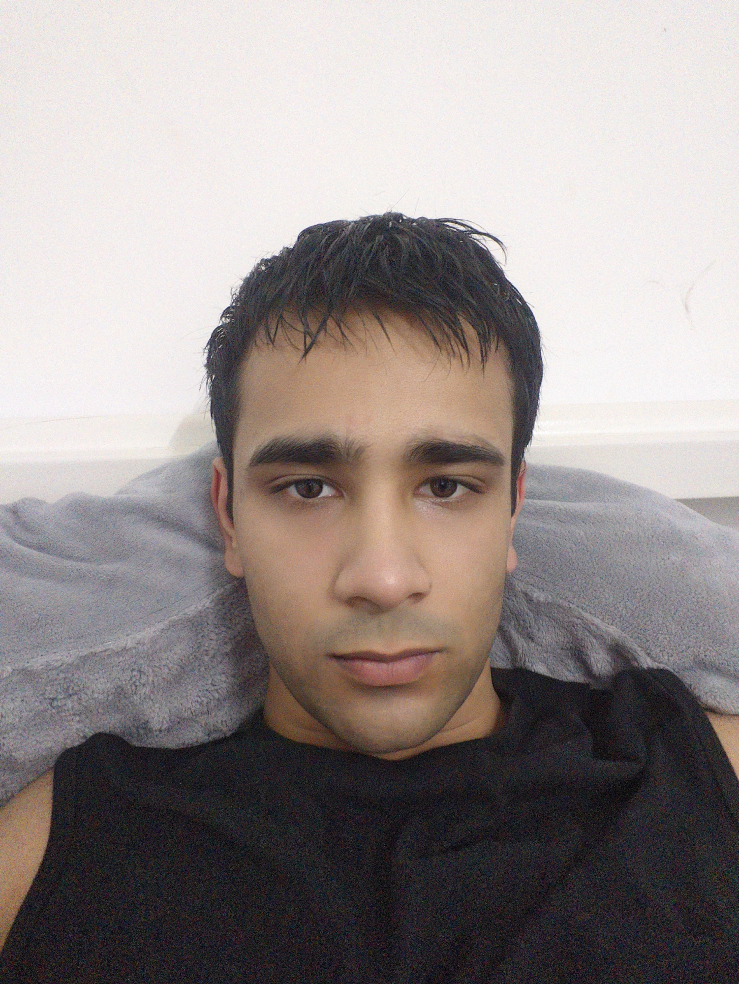 A close-up, slightly grainy, eye-level photograph shows a young man with dark hair and brown eyes. He is lying down against a grey pillow. He is wearing a black sleeveless shirt. The background is a plain white wall. The image does not provide any information that would link it to the city of Chongqing High-tech Zone, China.