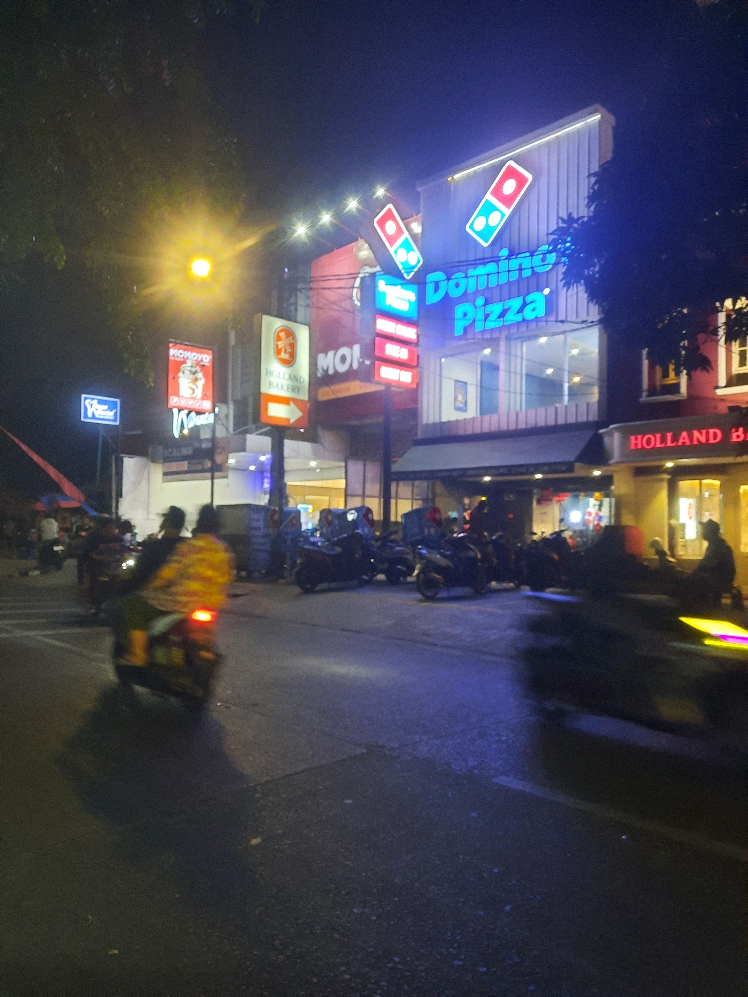 This is a nighttime, slightly blurry, full-frame, eye-level shot of a street scene in North Jakarta, Indonesia. The focus is on a Domino's Pizza restaurant with bright neon signage. Other illuminated signs for "Holland Bakery" and "Momoyo" are also visible. Several motorcycles are parked or moving on the street. Two people are riding a motorcycle in the foreground, with the passenger wearing a brightly colored jacket. The street is paved and appears wet in places, reflecting the neon lights. Trees are visible on the left and right sides of the image, with foliage silhouetted against the night sky. The overall atmosphere is that of a busy urban commercial area at night.