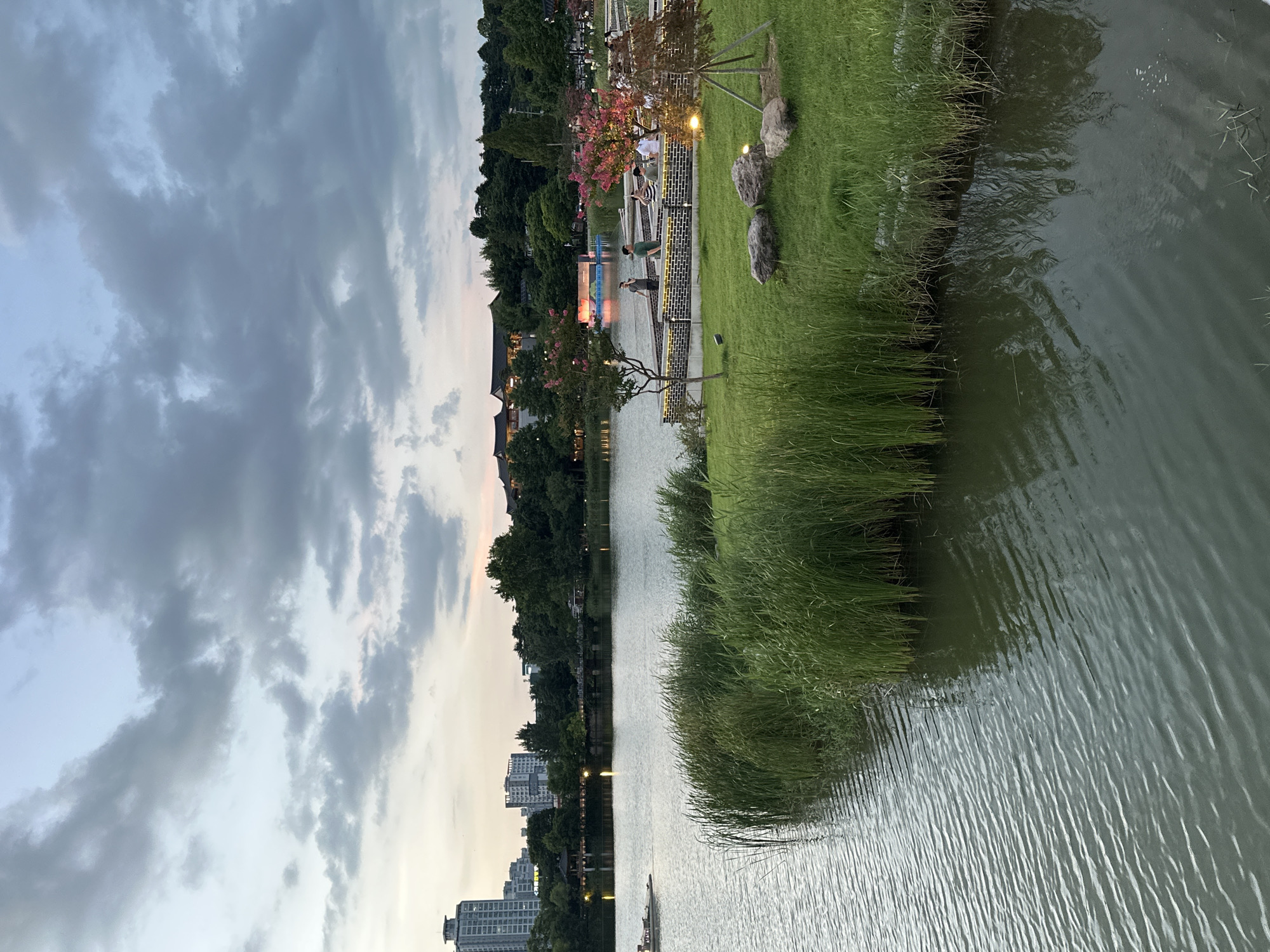 This is a photograph of a park with a lake, taken from a slightly elevated perspective on a cloudy evening. The foreground features a calm body of water with ripples, bordered by lush green grass and tall reeds. Beyond the water, a paved walkway runs along the edge of the lake, populated by several individuals. Some people are walking, while others are standing or sitting. A boat is visible on the left side of the lake, and in the background, there are trees, traditional-style buildings with dark roofs, and modern high-rise buildings. The sky is filled with a mix of gray and light blue clouds, suggesting the end of the day. The overall impression is a serene, urban park setting in Jeonju-si, South Korea.