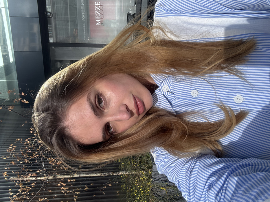 A female subject, appearing to be in her 20s or early 30s, is depicted in a close-up, outdoor selfie. She has long, light brown hair with darker roots and is wearing a blue and white striped button-down shirt. Her eyes are directed towards the camera. The scene is illuminated by direct sunlight, casting striped shadows across her shirt. In the background to the left, a dark, textured building facade is partially obscured by a tree with sparse, light-colored foliage. To the right, a modern glass-fronted building reflects the word "MEZZE" multiple times, indicating a restaurant. Outdoor seating with chairs and tables is visible through the reflection in the glass. Based on the "MEZZE" sign and architectural cues, the location is identified as being near **Shota Rustaveli Avenue 17 in Tbilisi, Georgia**, likely outside the Mezze restaurant.