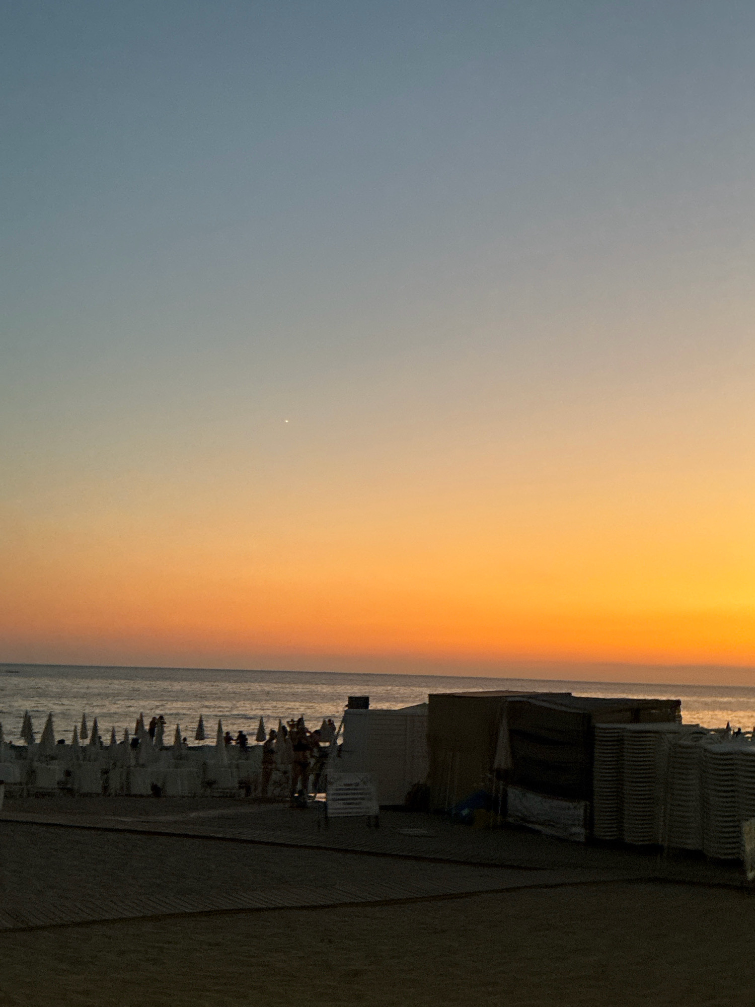 The image depicts a serene beach scene at dusk in Alanya, Turkey. The sky is a gradient of pale blue at the top, transitioning to a vibrant orange and yellow near the horizon, indicating the setting sun. A single bright star is visible in the upper portion of the sky. The sea is calm, with gentle waves meeting the shore.
In the foreground, a sandy beach leads to the water. A wooden boardwalk or pathway is visible on the sand. Further back, rows of white beach chairs and umbrellas are neatly arranged, suggesting a resort or public beach area. Some people are visible in the distance, either walking or relaxing on the beach.
To the right of the image, there is a structure with a dark tarp covering it, and stacked chairs are piled beside it. A few smaller structures are also present along the beach. The overall atmosphere is tranquil and peaceful, capturing the end of a day at the beach.