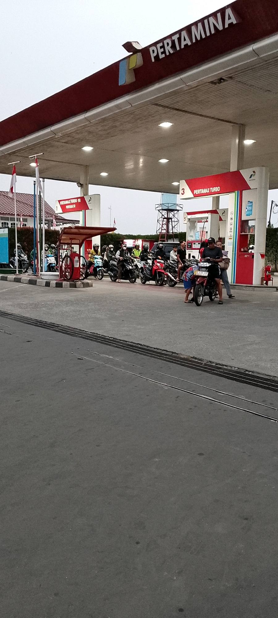 The image depicts a gas station in Depok, Indonesia, specifically identifiable as a Pertamina station due to the prominent branding. The scene is filled with activity, primarily centered around motorcycles. Several motorcycles are lined up, appearing to be waiting to refuel, with some riders seated on their bikes. One motorcycle in the foreground has two individuals associated with it. One person is crouched down, possibly attending to the motorcycle, while another stands beside them. In the background, more motorcycles and individuals are visible, suggesting a busy period at the gas station. The station itself features red and white signage, typical of Pertamina, with "PERTAMAX TURBO" prominently displayed on a pump. The overall environment suggests an outdoor, public space with overhead lighting visible on the station canopy. The ground is paved, and a set of parallel metal grates or tracks are visible in the lower portion of the image. The sky appears overcast.